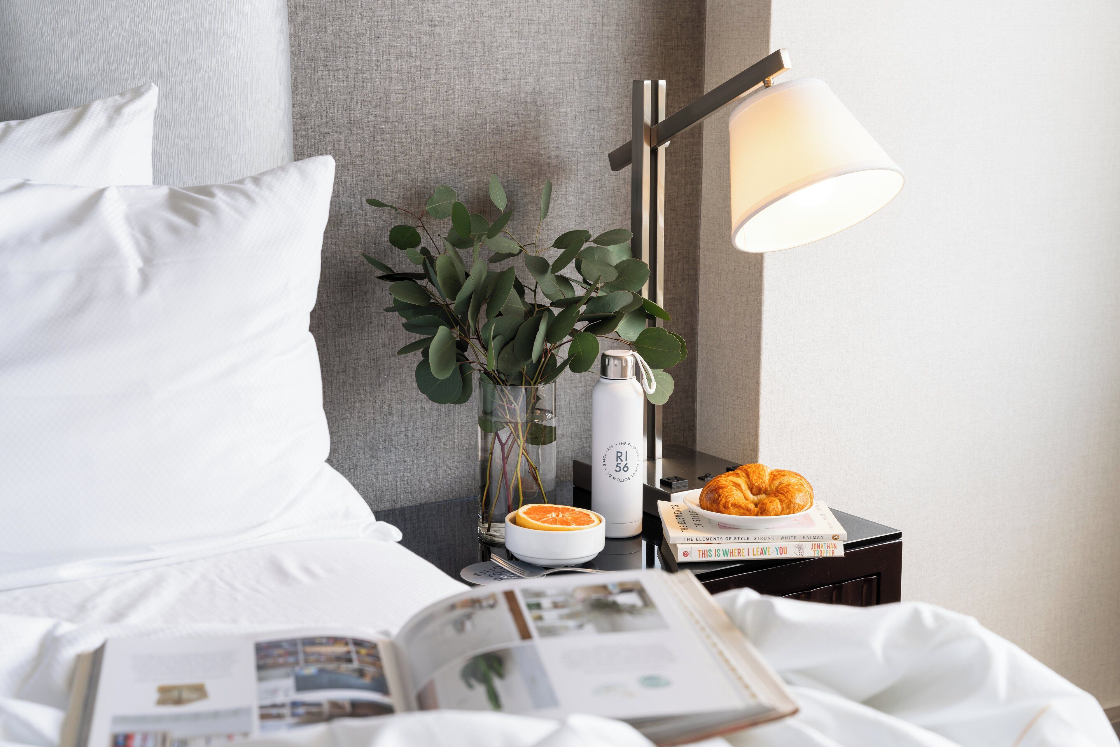 Een net opgemaakt bed met witte lakens, een nachtkastje met een lamp, een boek, een croissant, een glazen vaas met groene bladeren en een waterfles.
