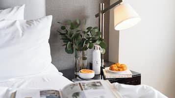 Een net opgemaakt bed met witte lakens, een nachtkastje met een lamp, een boek, een croissant, een glazen vaas met groene bladeren en een waterfles.
