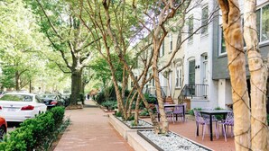 Una acera bordeada de árboles con coches estacionados, mesas al aire libre y un edificio con múltiples ventanas y balcones.