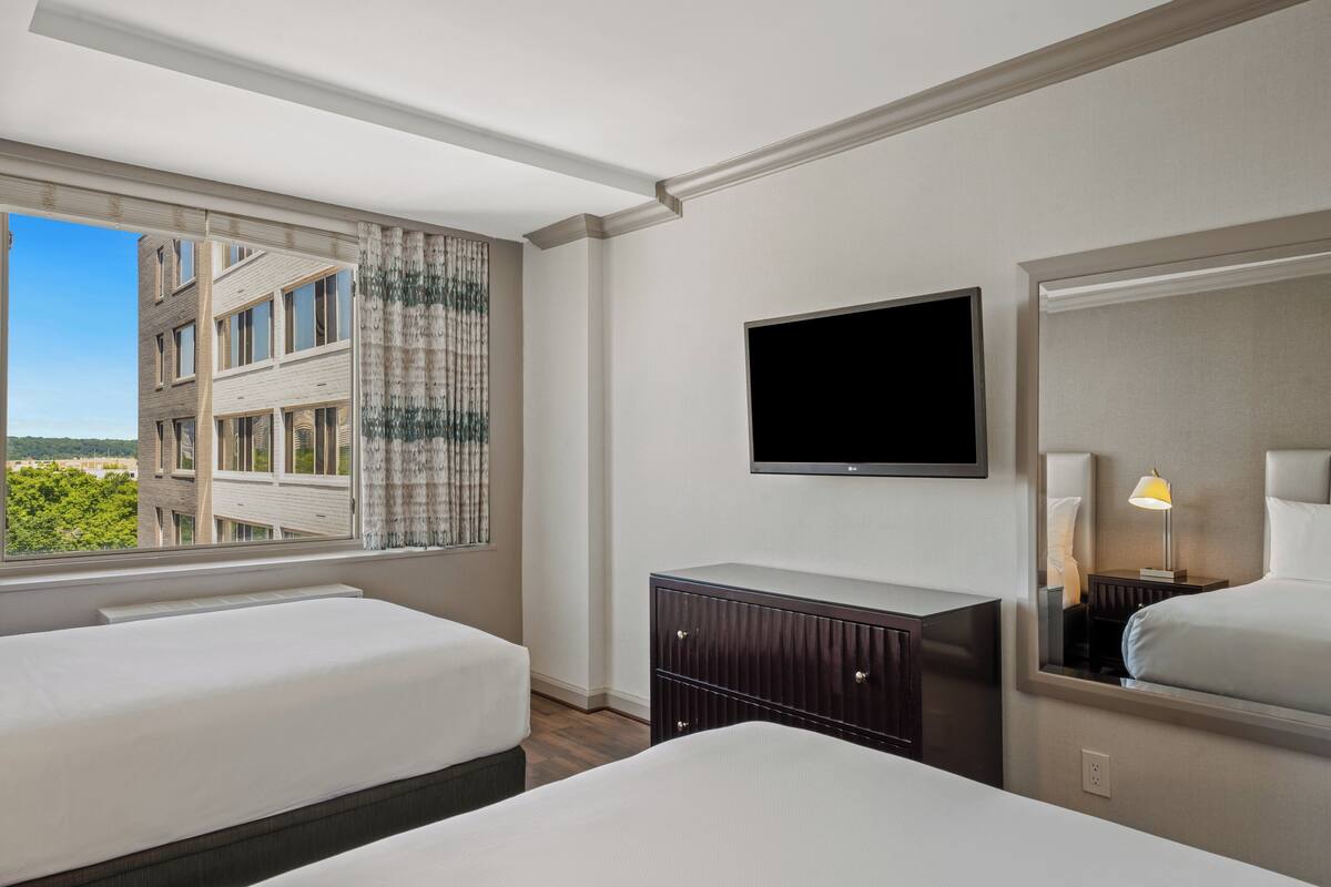A hotel room with two beds, a TV, a dresser, and a window with a view of buildings.