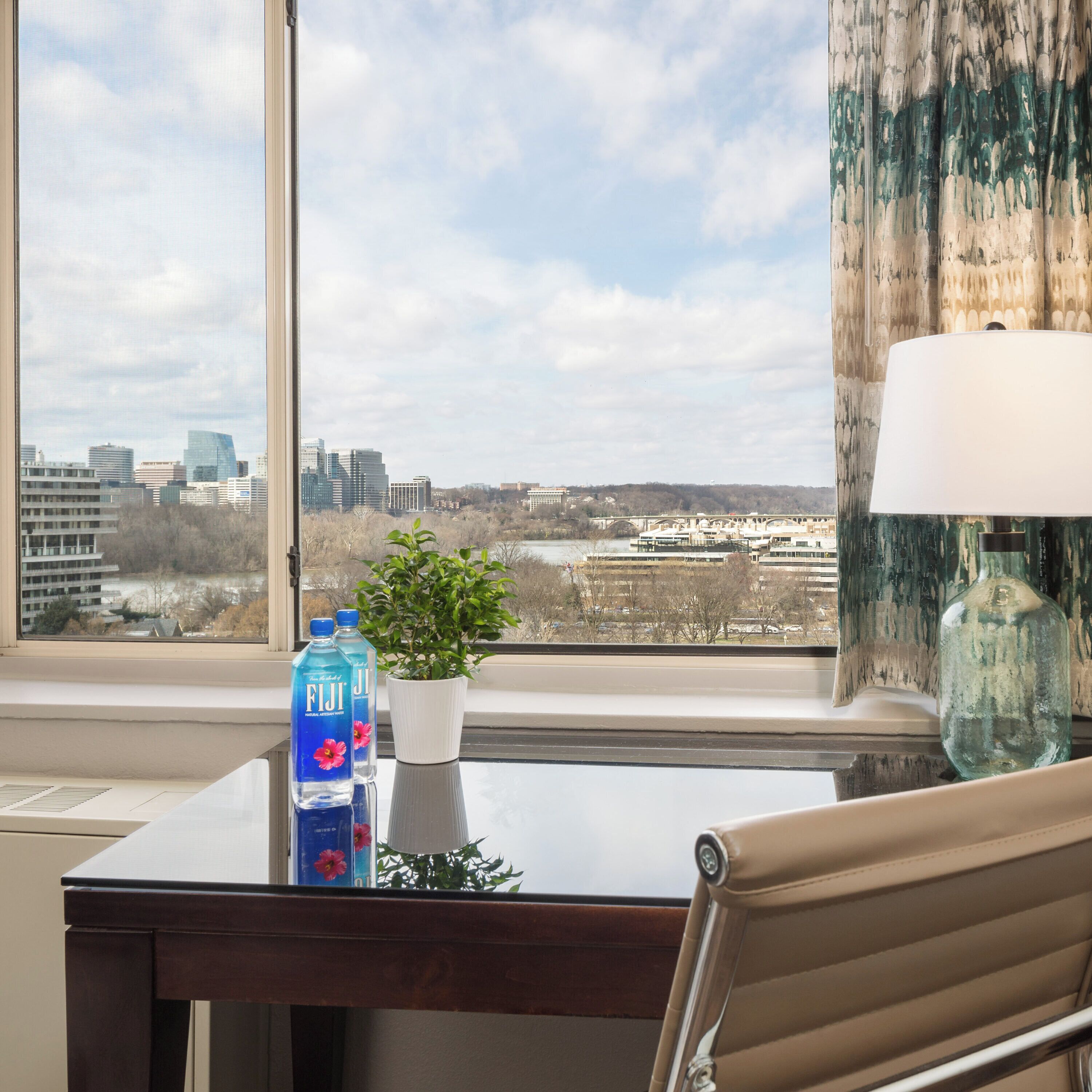 A hotel room with a view of a cityscape, a desk with a lamp, a potted plant, and two bottles of Fiji water.