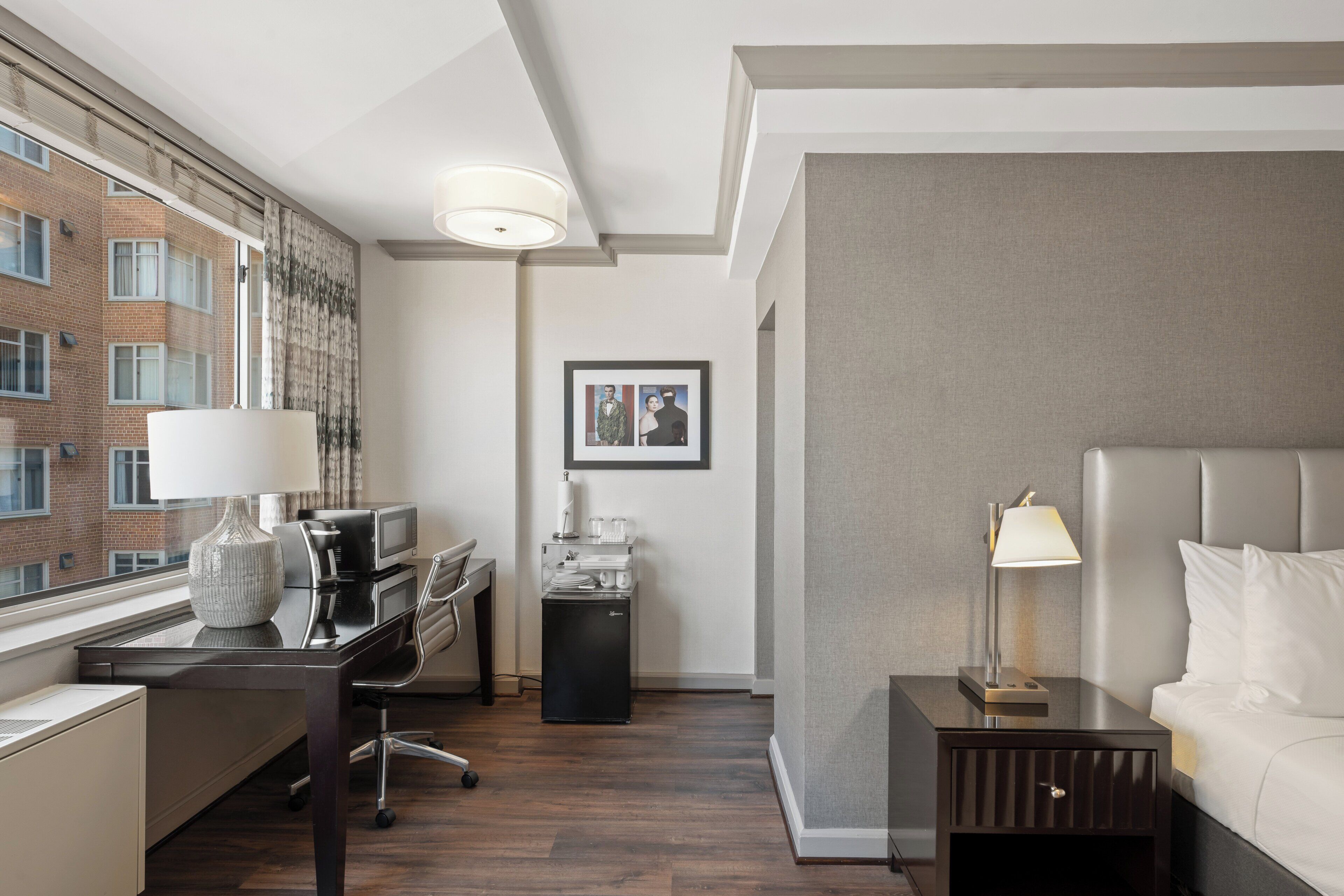 A hotel room with a bed, desk, and chair, a small kitchenette, and a window with a view of a brick building.