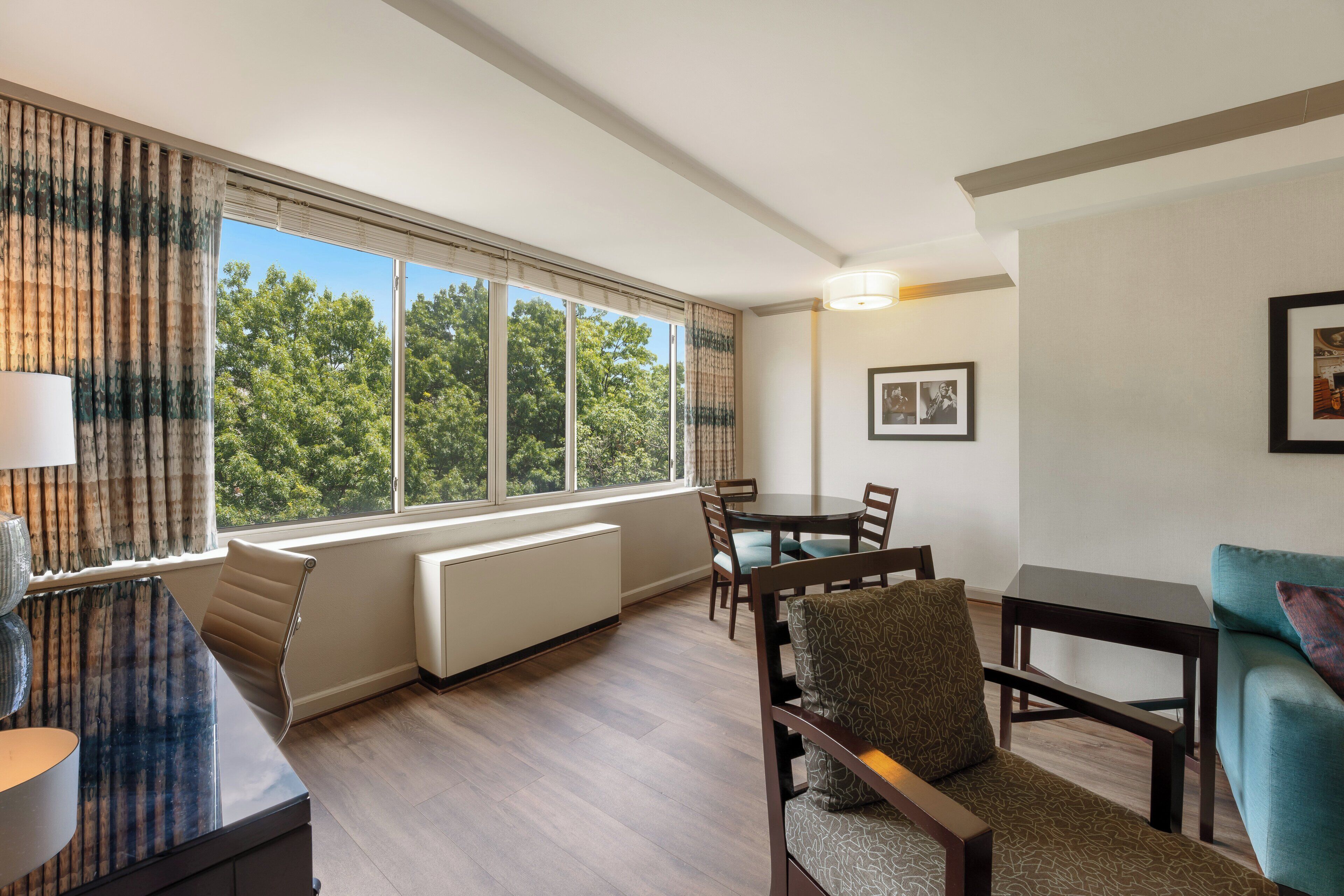 A modern hotel room with a dining area, large windows with a view of trees, a radiator, and a lamp.