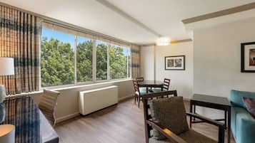 A modern hotel room with a dining area, large windows with a view of trees, a radiator, and a lamp.