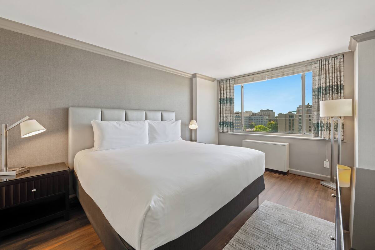A hotel room with a large bed, a nightstand, a lamp, a window with a view of buildings, and a radiator.