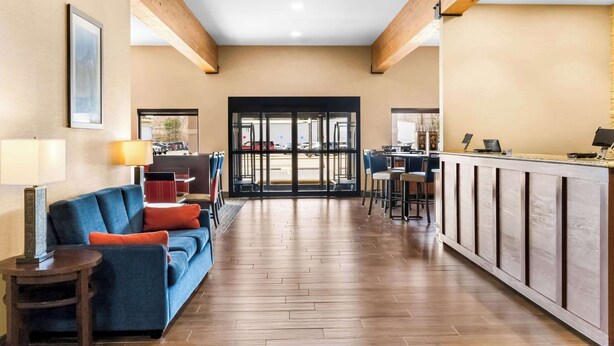Un hall d’hôtel avec un plancher en bois, un canapé bleu agrémenté de coussins orange, un comptoir de réception et une vue sur le parking, visible à travers des portes vitrées.