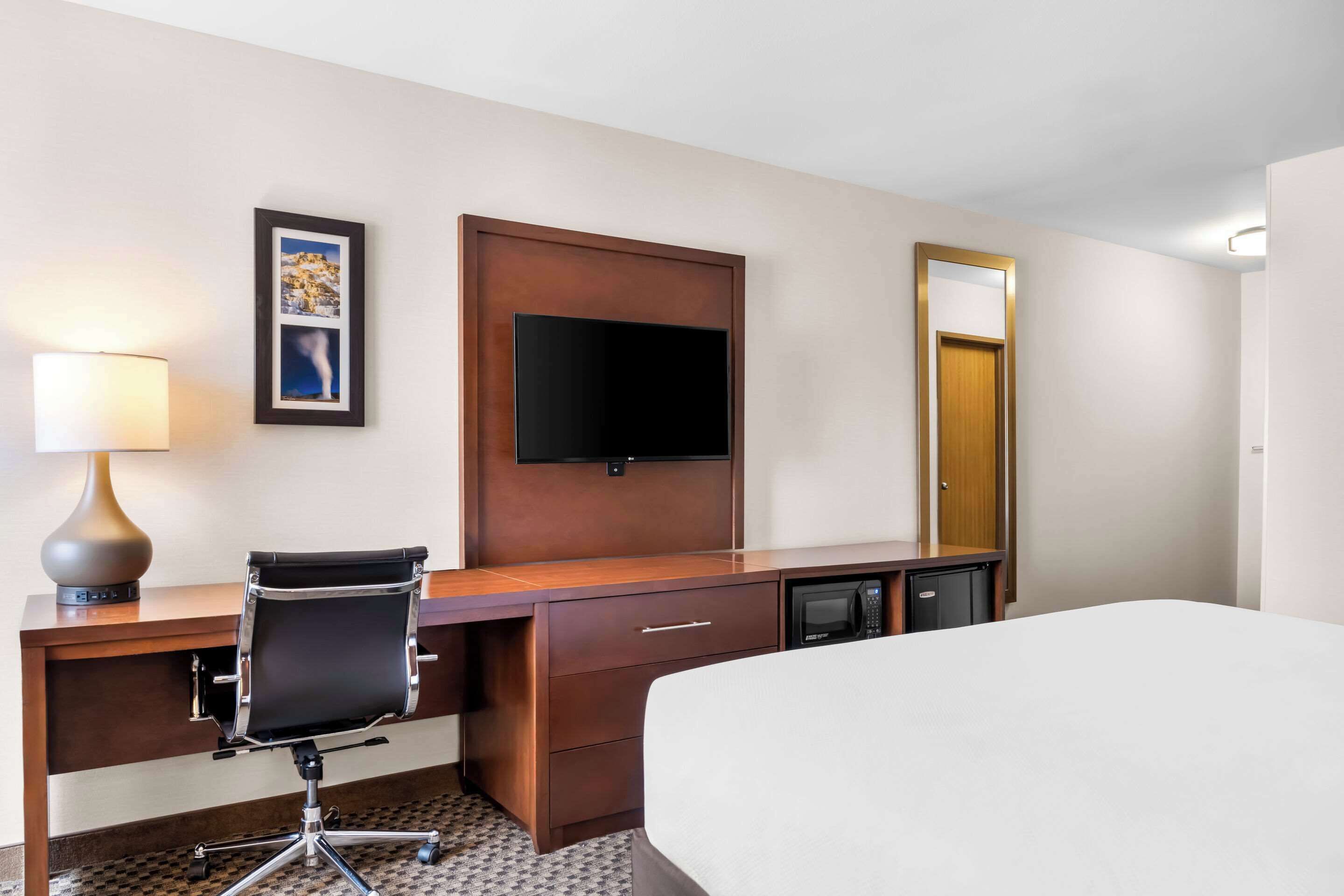A hotel room with a bed, a desk with a chair, a television, a microwave, and a door with a mirror.