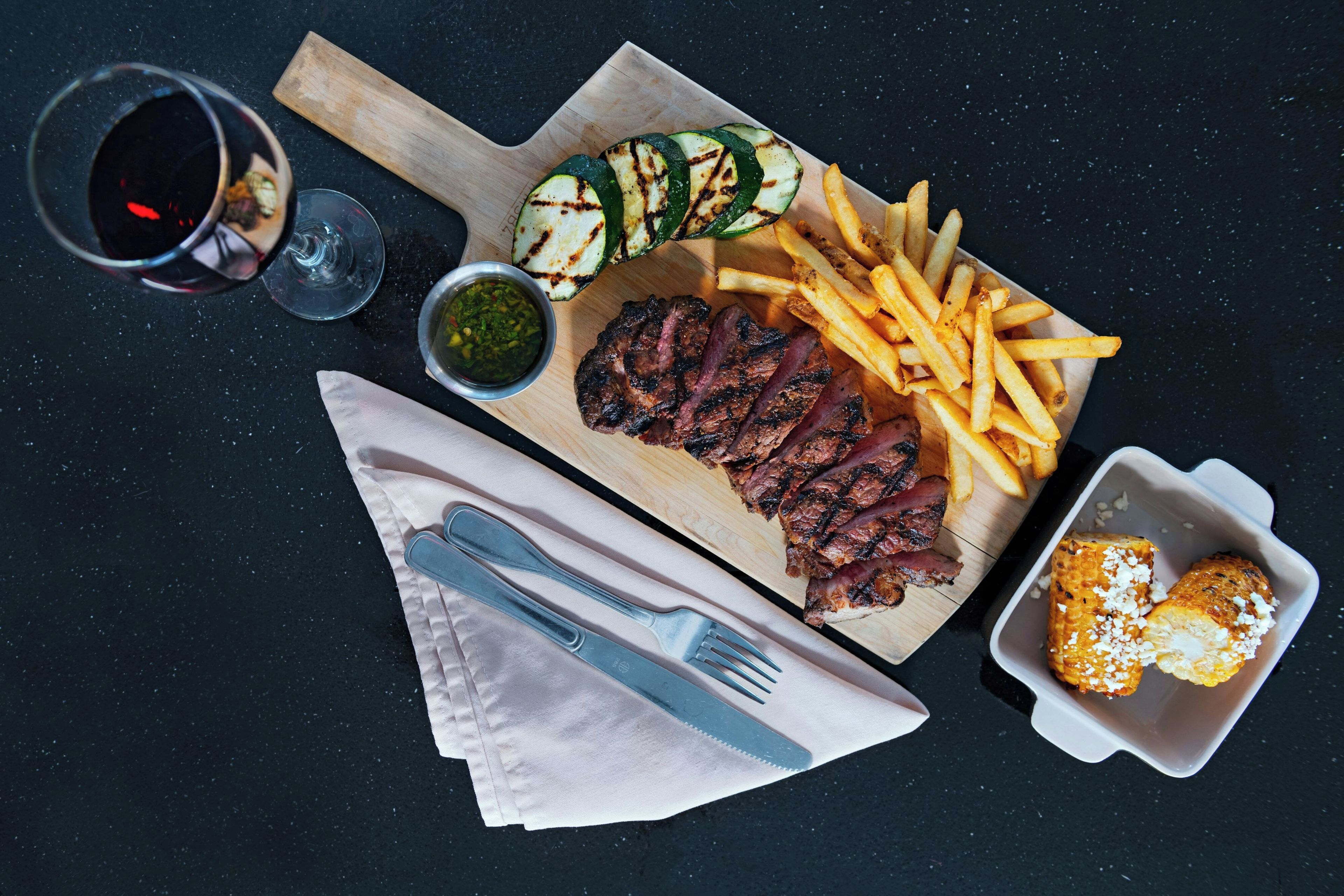 A meal with steak, fries, grilled vegetables, and corn on the cob on a wooden board.