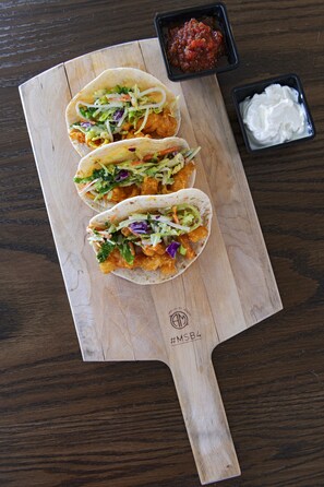 Tres tacos sobre una tabla de madera, con dos recipientes pequeños de salsa y una cucharada de crema agria.