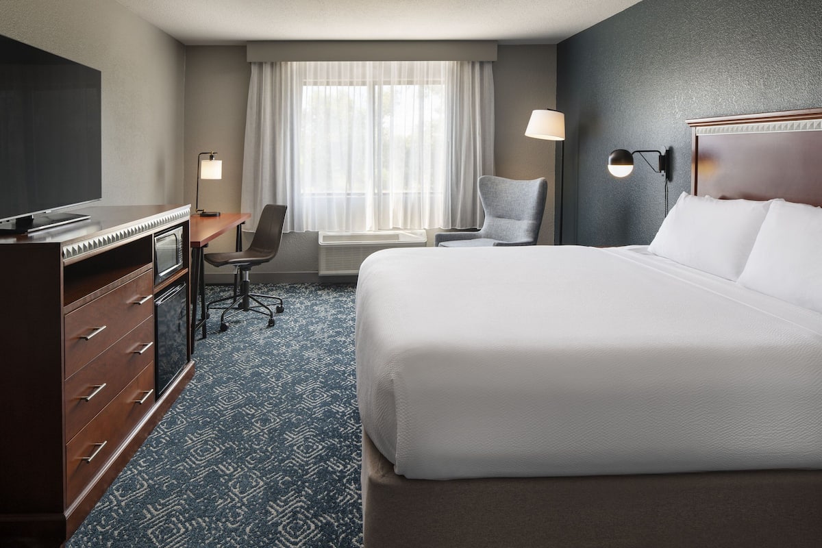 Ein Hotelzimmer mit einem großen Bett, einem Schreibtisch mit Stuhl, einem Fernseher und einem Fenster mit Vorhängen.