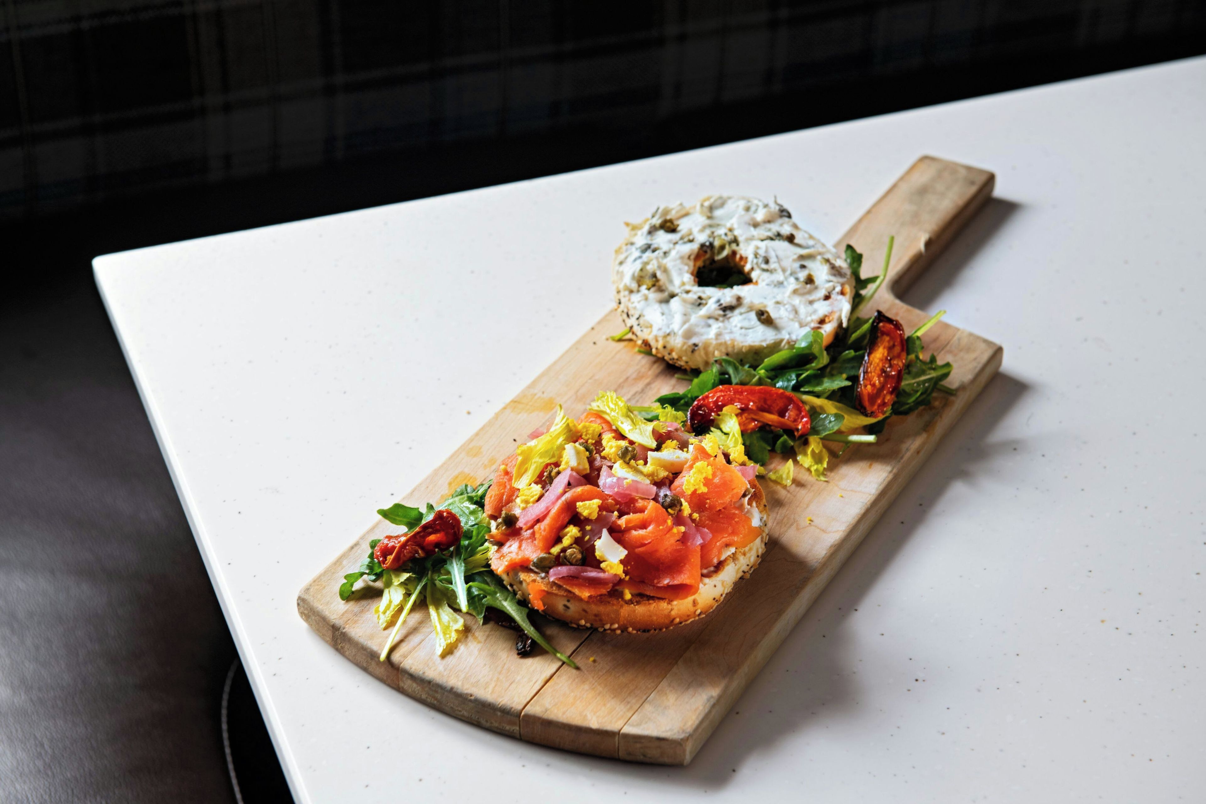 Un tagliere in legno con un bagel farcito con salmone affumicato, capperi e insalata, e un bagel semplice su una superficie bianca.