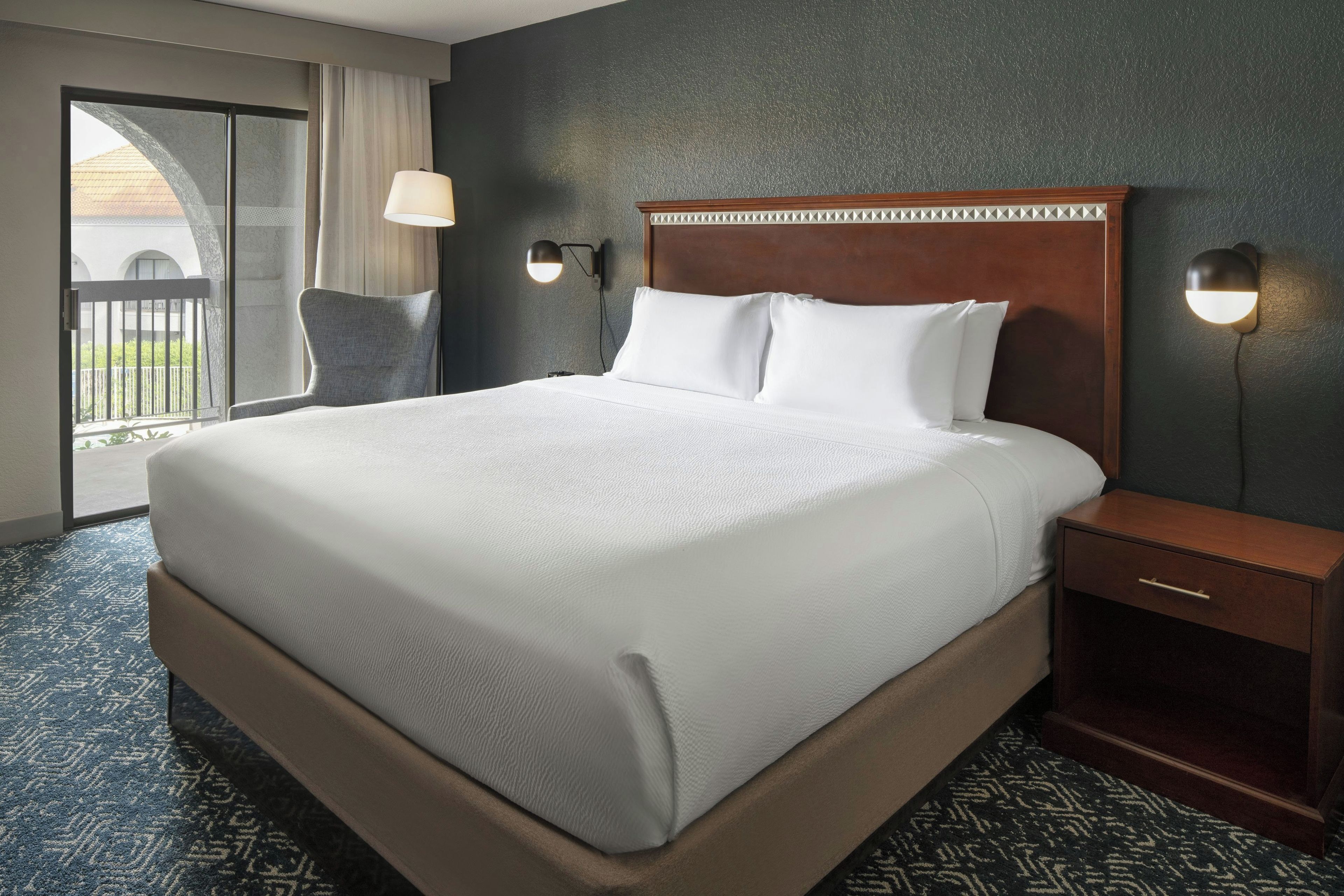 Une chambre d’hôtel avec un grand lit, une table de chevet, une chaise, une lampe fixée au mur et une vue sur l’extérieur.