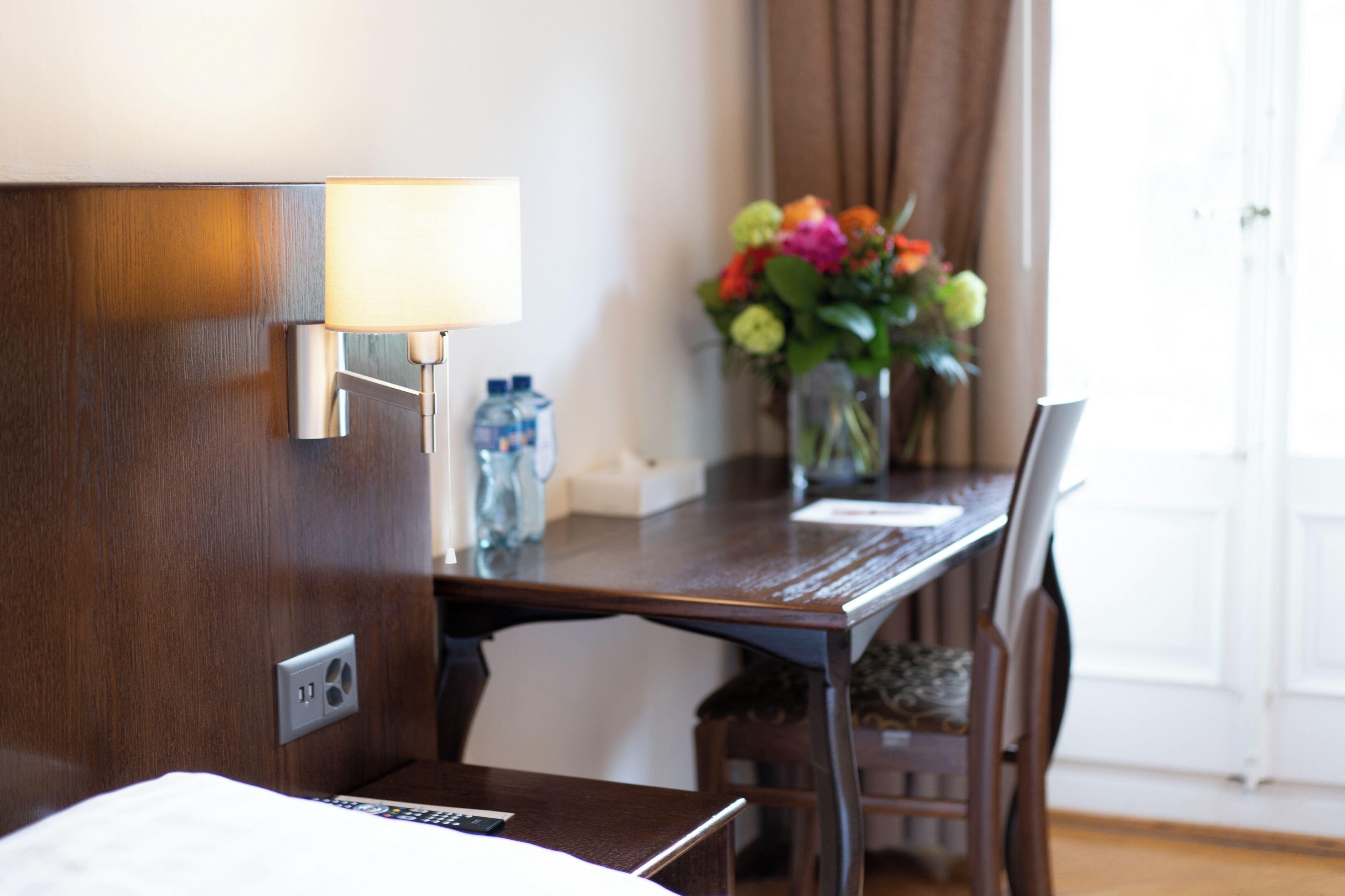 Habitación de hotel con un escritorio de madera, una lámpara, un florero con flores y una ventana con cortinas.