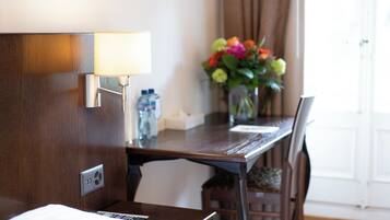 Habitación de hotel con un escritorio de madera, una lámpara, un florero con flores y una ventana con cortinas.