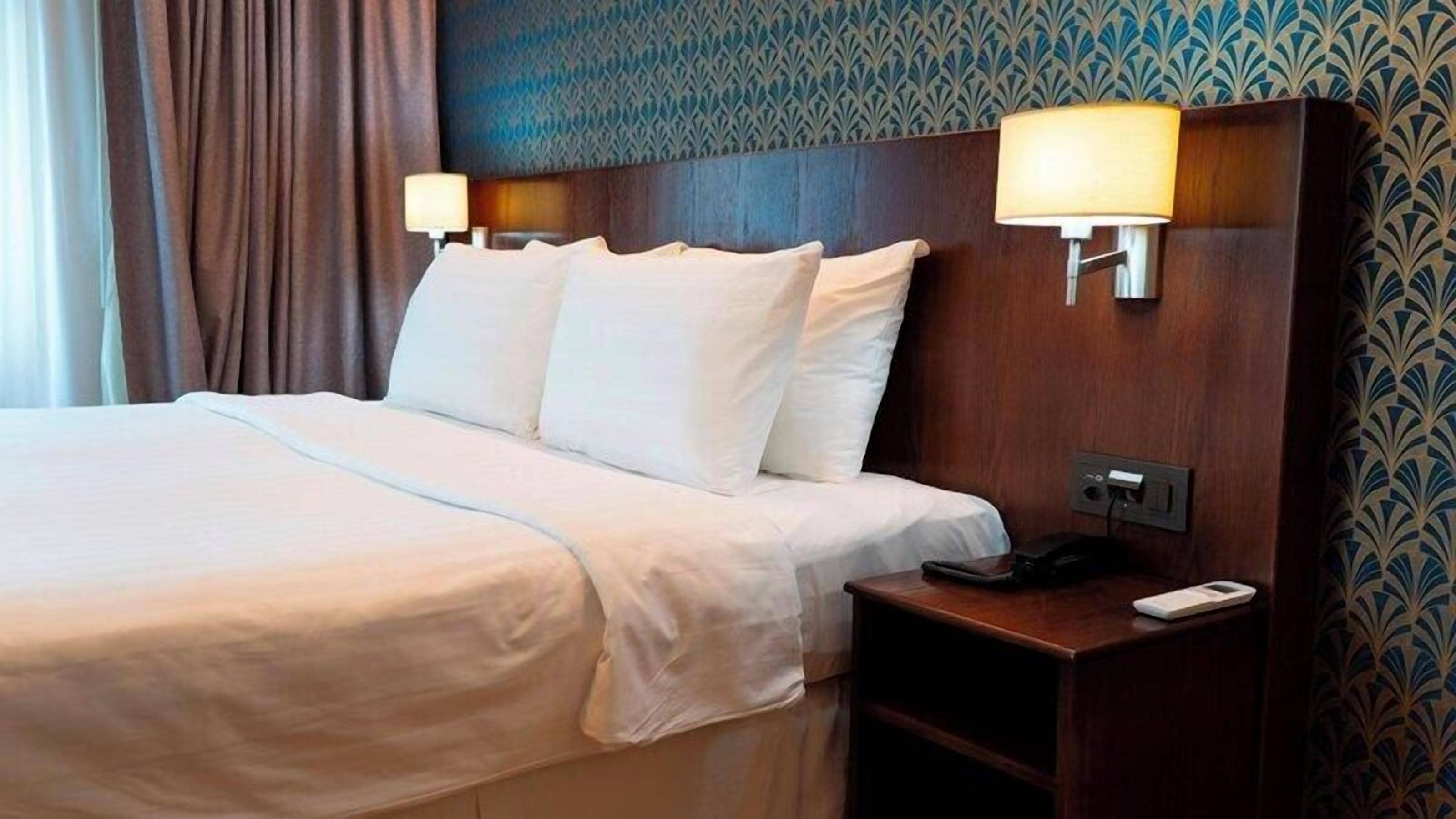 A hotel room with a large bed, two bedside lamps, a nightstand, and a patterned wallpaper.