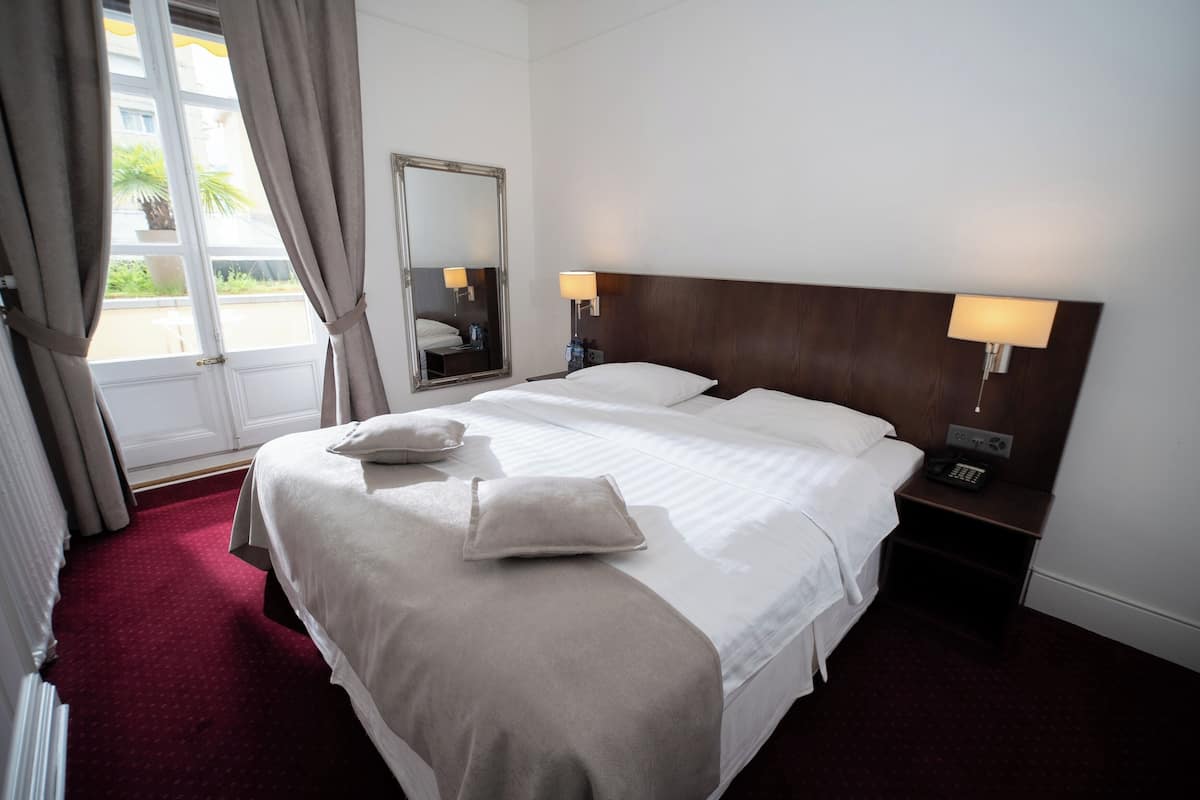 Ein Hotelzimmer mit einem großen Bett, zwei Nachttischen mit Lampen, einem Spiegel und Blick ins Freie durch ein Fenster.