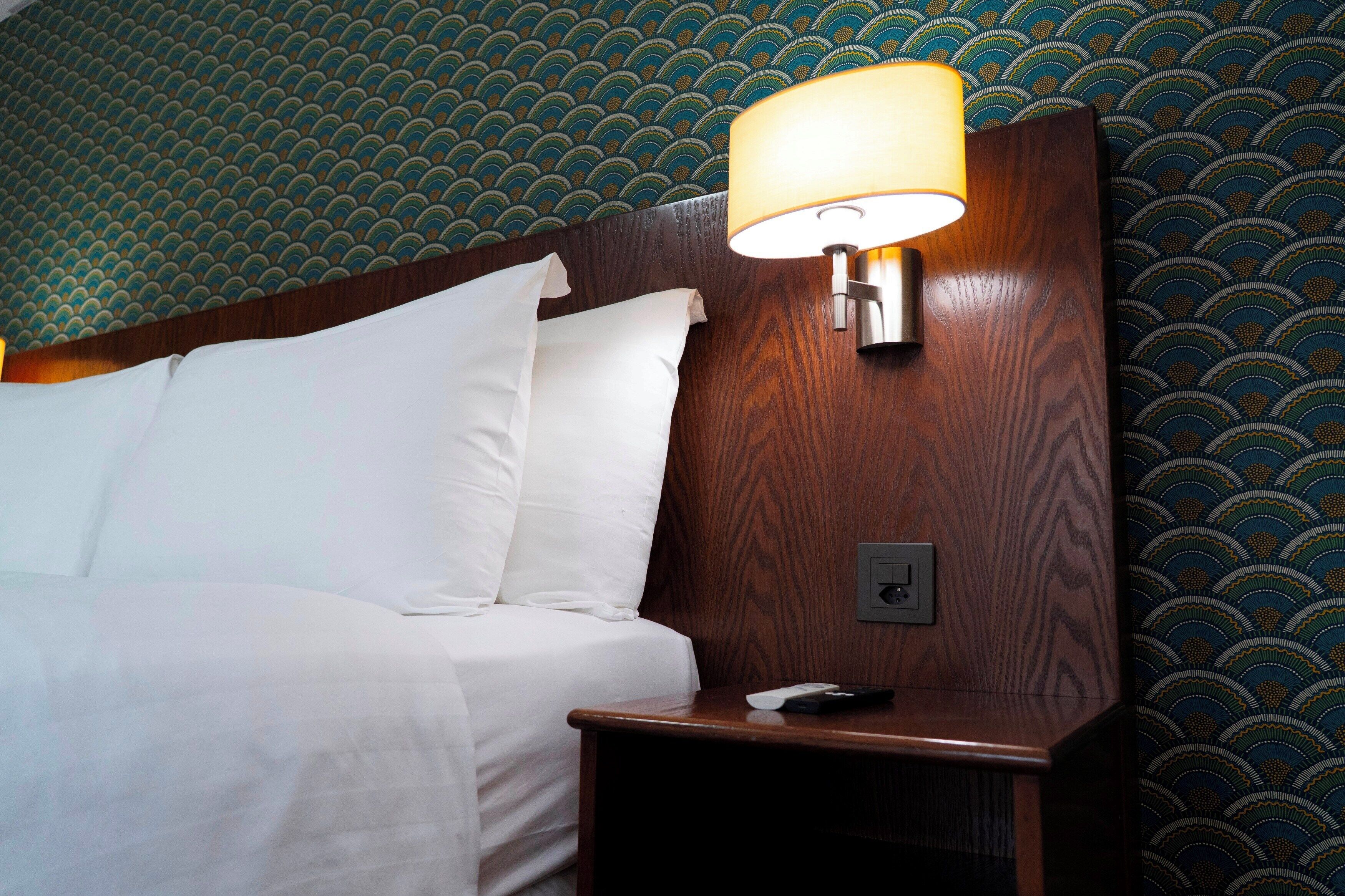 A hotel room with a wooden headboard, white bedding, a bedside lamp, and a patterned wallpaper.