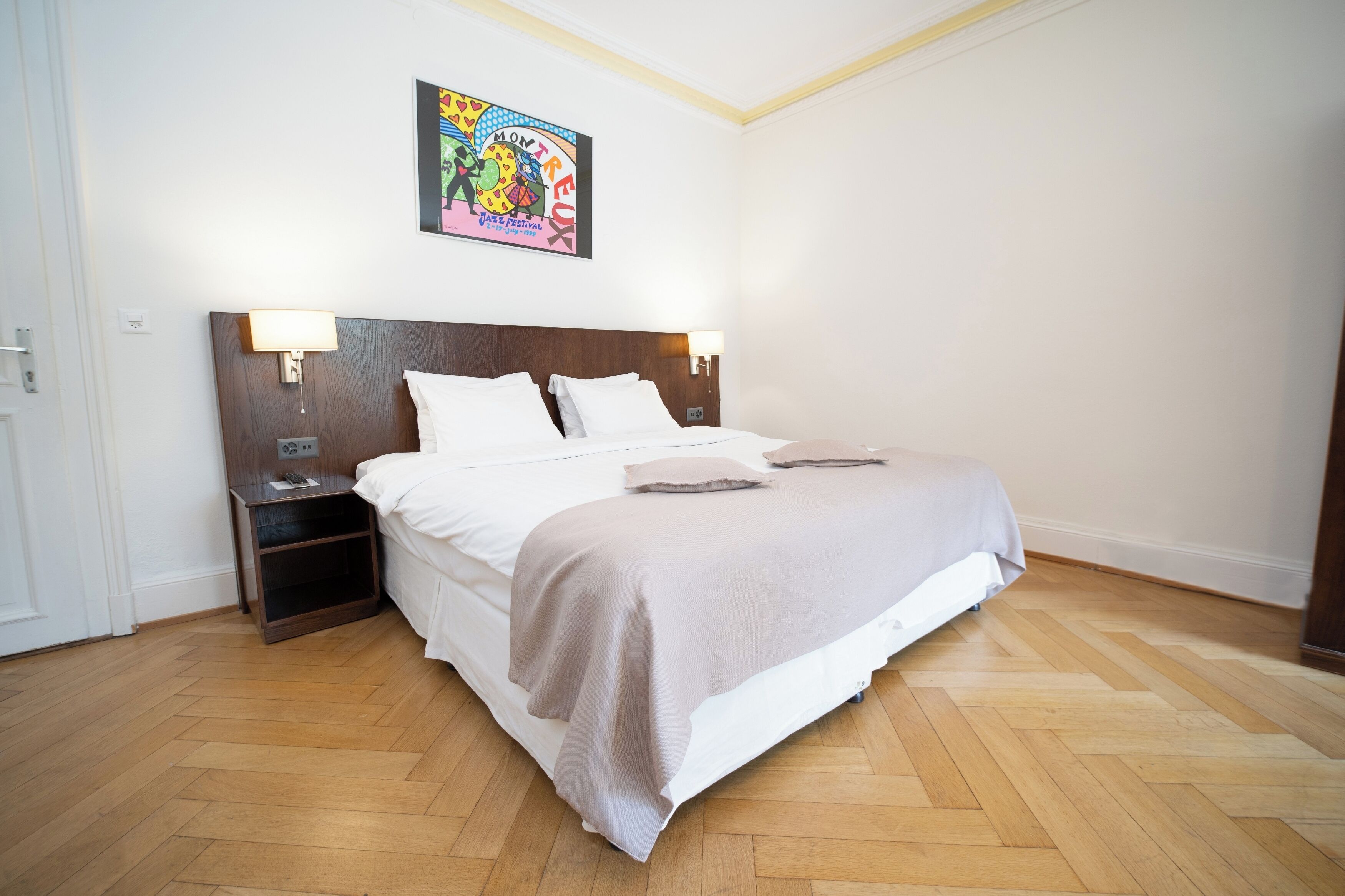 Ein ordentlich bezogenes Bett mit einem hölzernen Kopfteil, zwei Nachttischen mit Lampen und einem farbenfrohen Bild an der Wand.