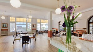 A spacious dining area with round tables and chairs, large windows offering a view of the city, and a vase with flowers on the counter. - J5 Hotels Helvetie Montreux (Montreux)