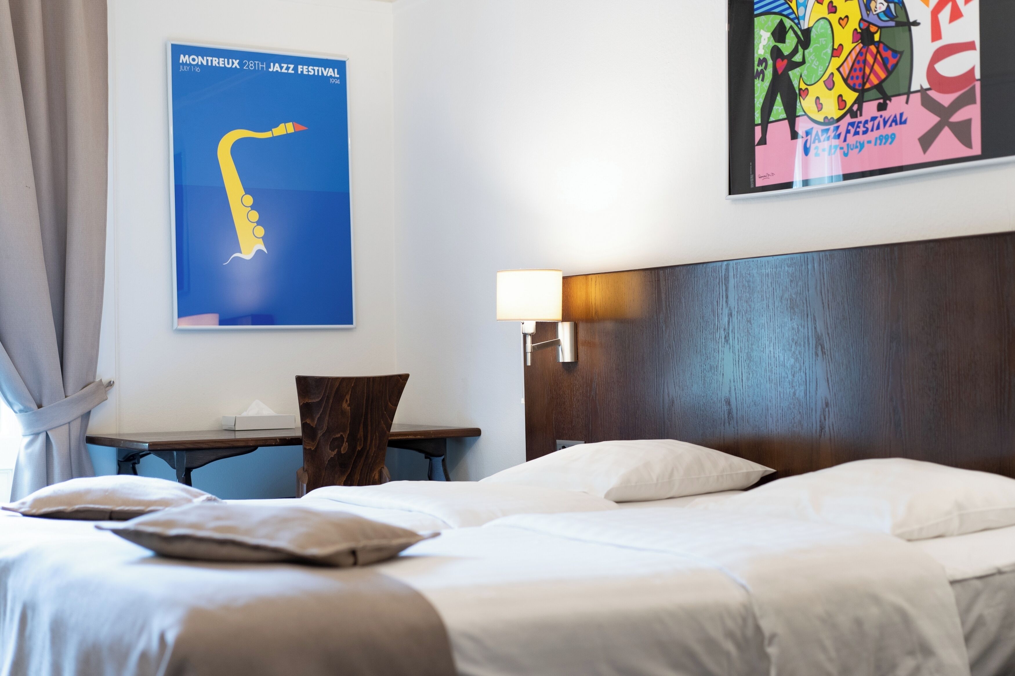 A hotel room with a large bed, a desk, a chair, and two posters on the wall.