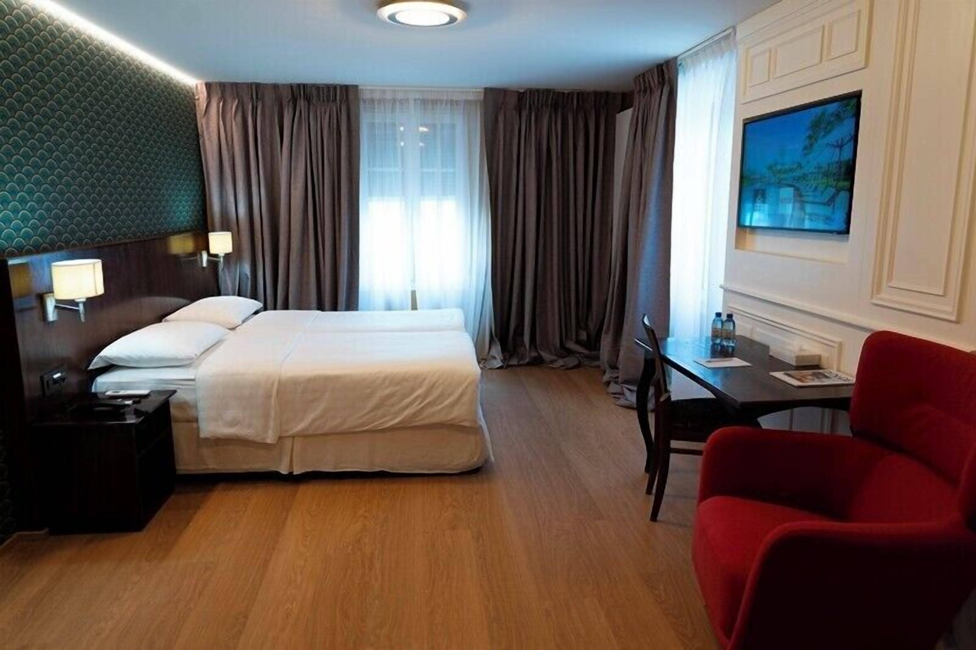 A hotel room with a double bed, a desk with a chair, a TV mounted on the wall, and a red armchair.