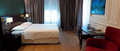 A hotel room with a double bed, a desk with a chair, a TV mounted on the wall, and a red armchair.