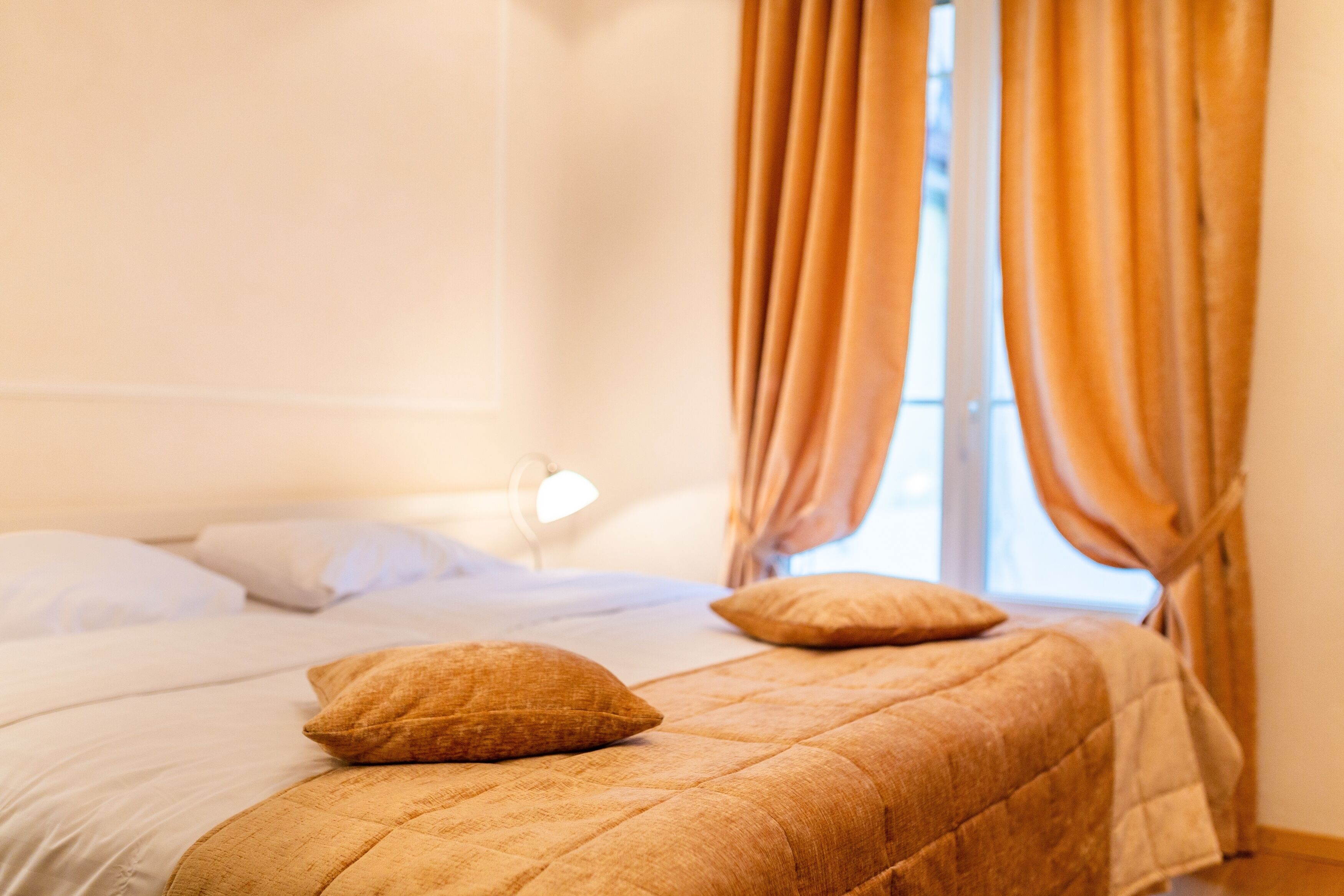 A neatly made bed with a quilted cover and two pillows, a bedside lamp, and curtains on a window.