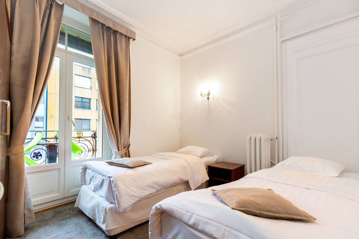 Ein Hotelzimmer mit zwei Betten, einem Fenster mit Vorhängen, einem Heizkörper und einem Nachttisch.