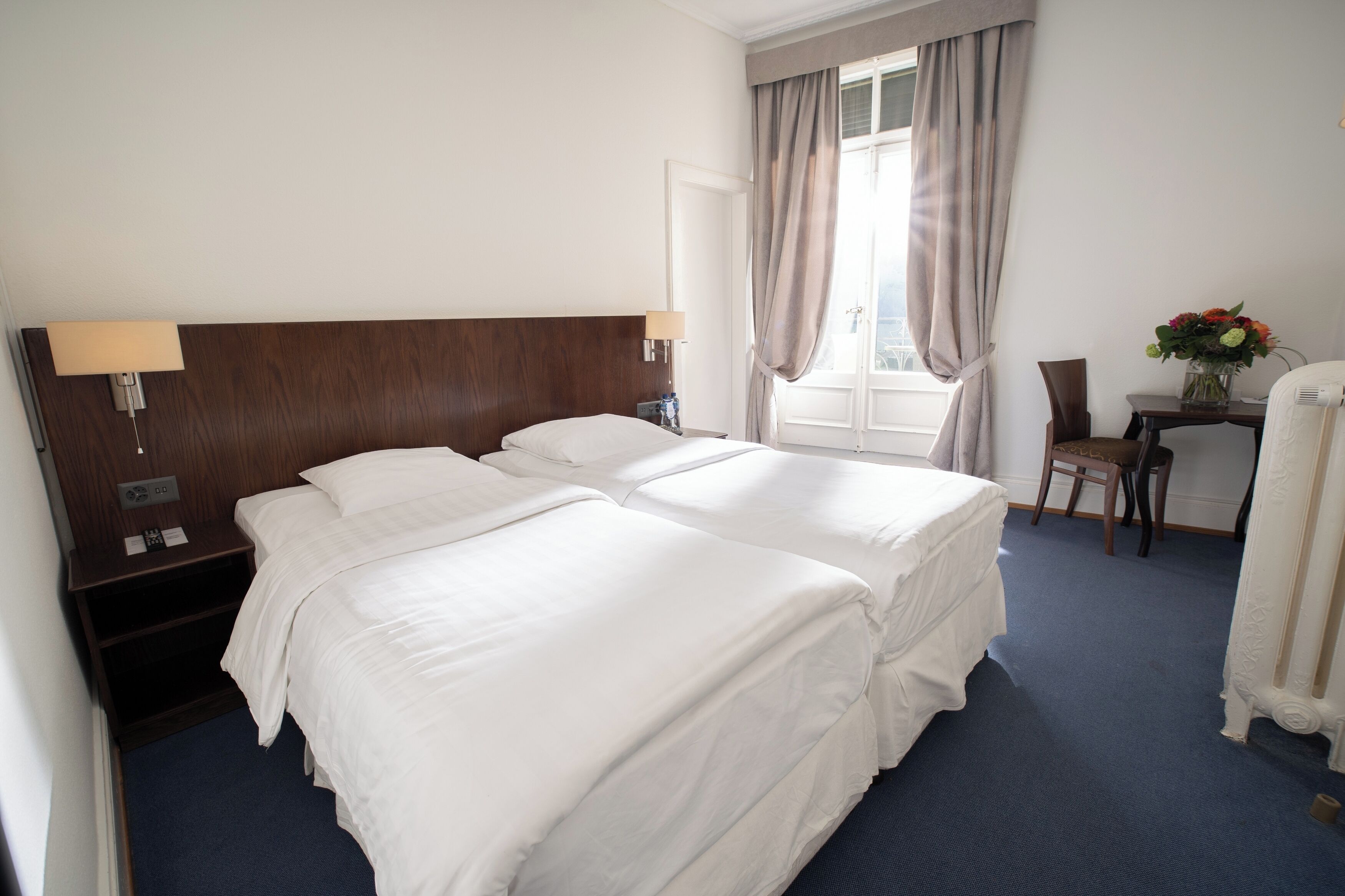 Habitación de hotel con dos camas, un escritorio, una silla, una lámpara y un ventanal con cortinas.