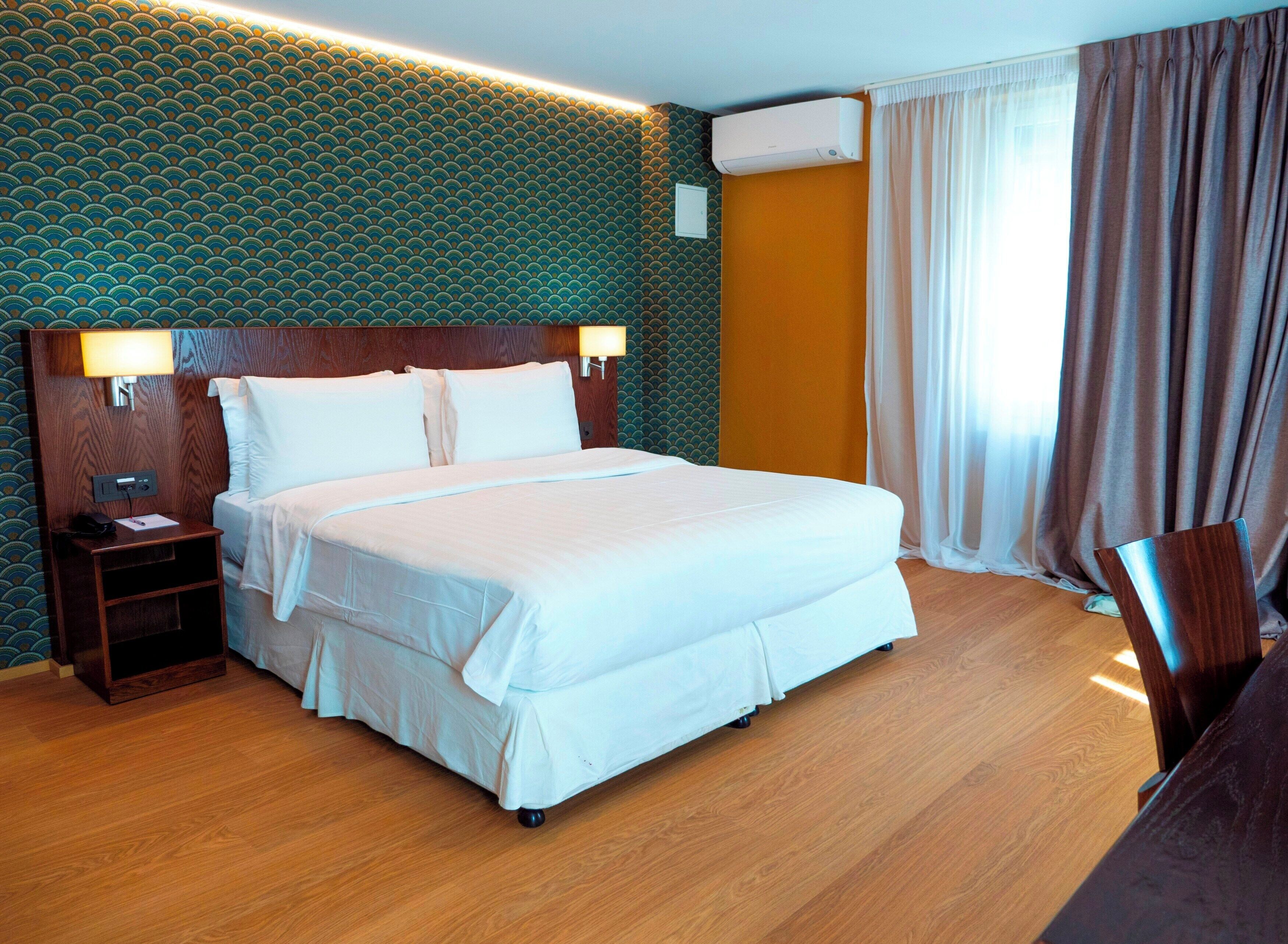 A hotel room with a large bed, a wooden nightstand, a wall-mounted air conditioner, and a window with curtains.