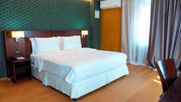 A hotel room with a large bed, a wooden nightstand, a wall-mounted air conditioner, and a window with curtains.
