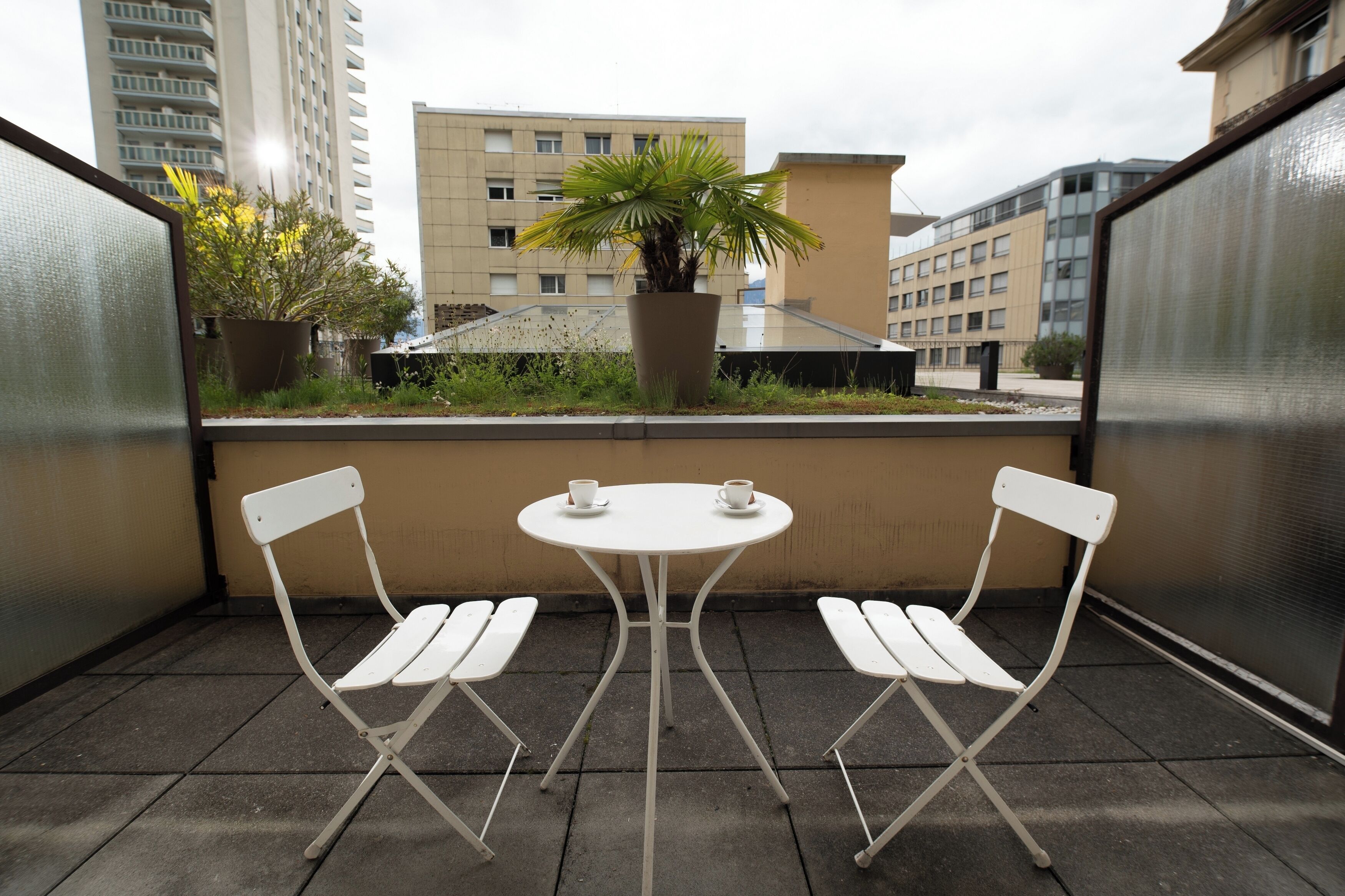 A balcony with two white folding chairs and a small round table, a potted plant, and a view of buildings in the background.