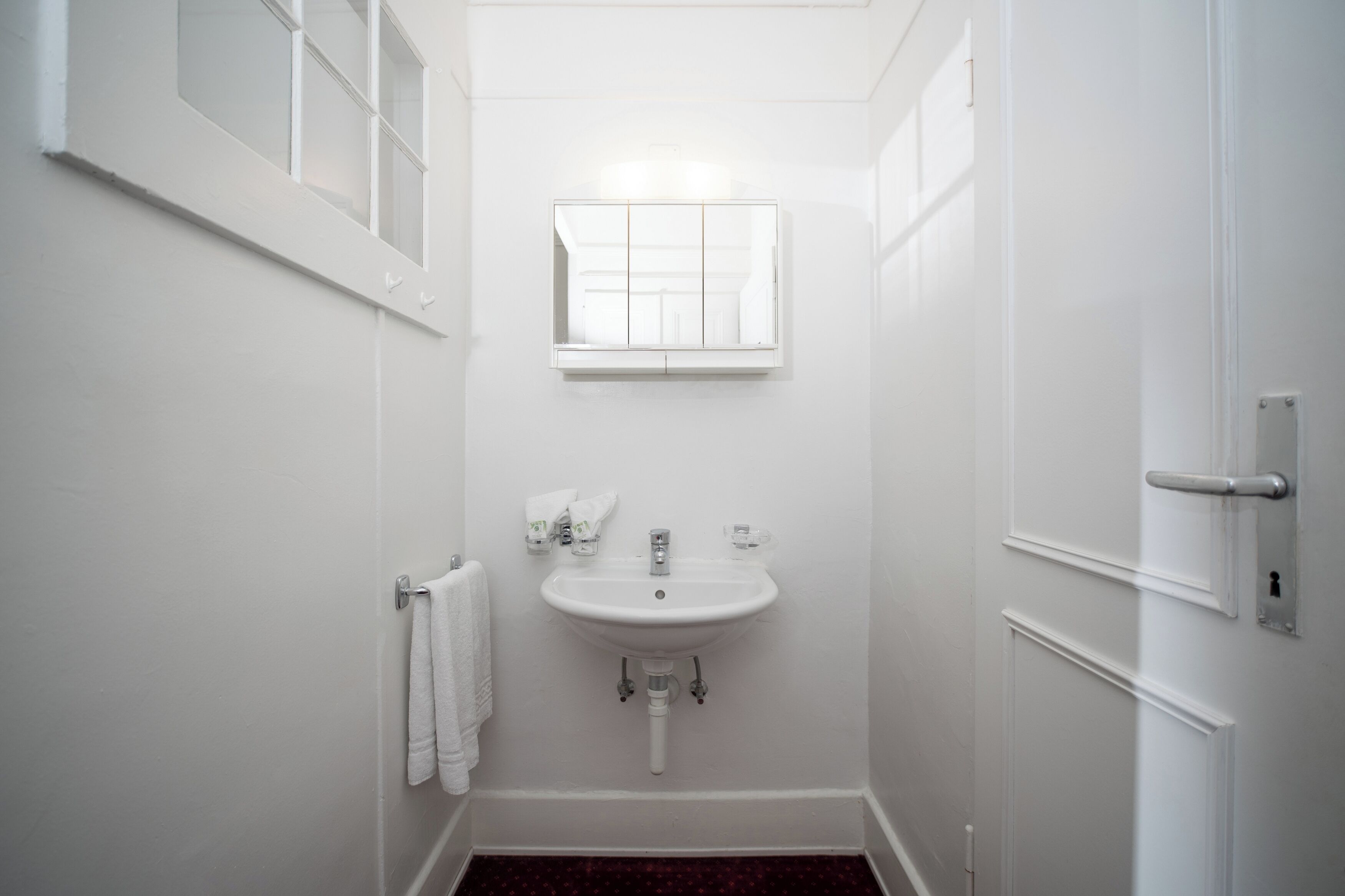 Ein kleines, weißes Badezimmer mit einem Standwaschbecken, einem Spiegel darüber und einem Handtuch an der Wand.