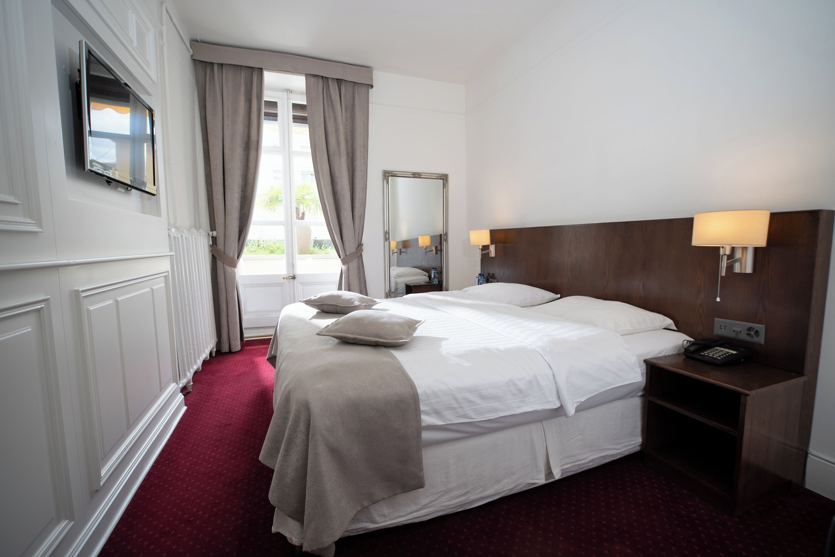A hotel room with a large bed, a TV mounted on the wall, a bedside table with a lamp, and a window with curtains.
