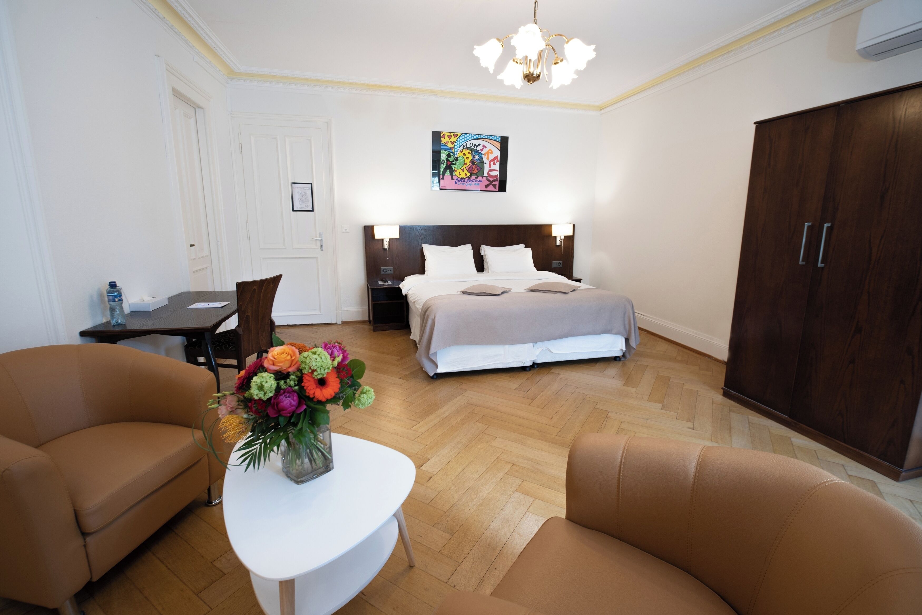 A hotel room with a bed, a desk, two chairs, a small table with a vase of flowers, a wardrobe, and a ceiling light.