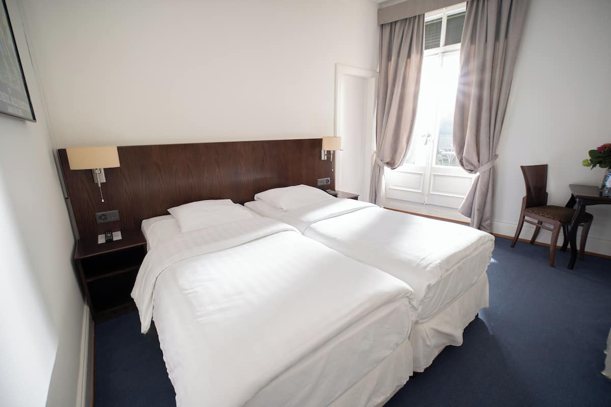 Ein Hotelzimmer mit zwei Betten, einem Schreibtisch, einem Stuhl, einem Fenster mit Vorhängen und einer Wandlampe.