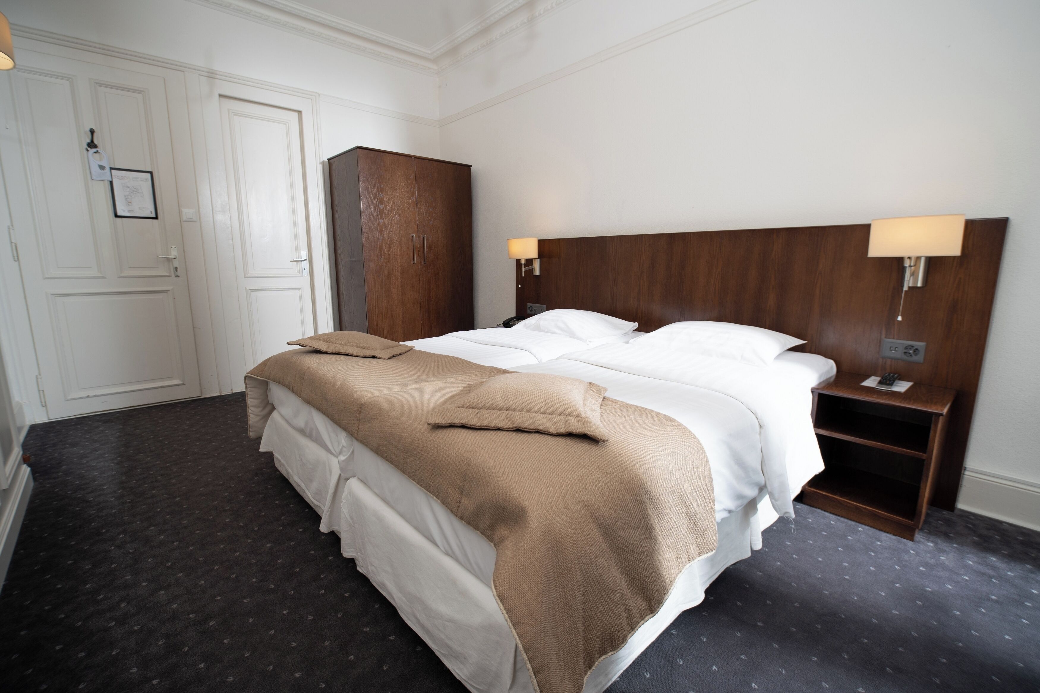 Una habitación de hotel con una cama grande, dos mesitas de noche, un armario y una puerta con un cuadro enmarcado.