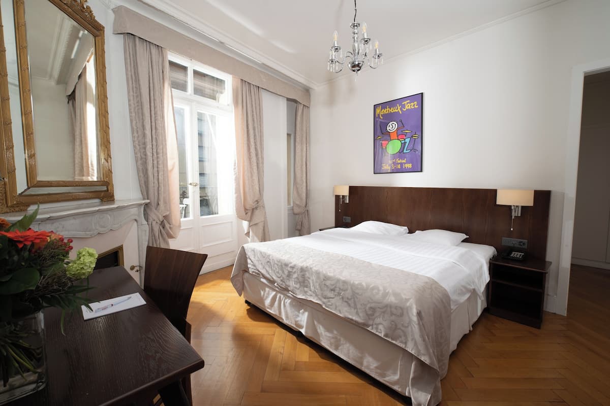 Ein Hotelzimmer mit einem großen Bett, einem Holfnachtkästchen, einem Schreibtisch mit Blumenvase, einem Spiegel und einem Poster an der Wand.