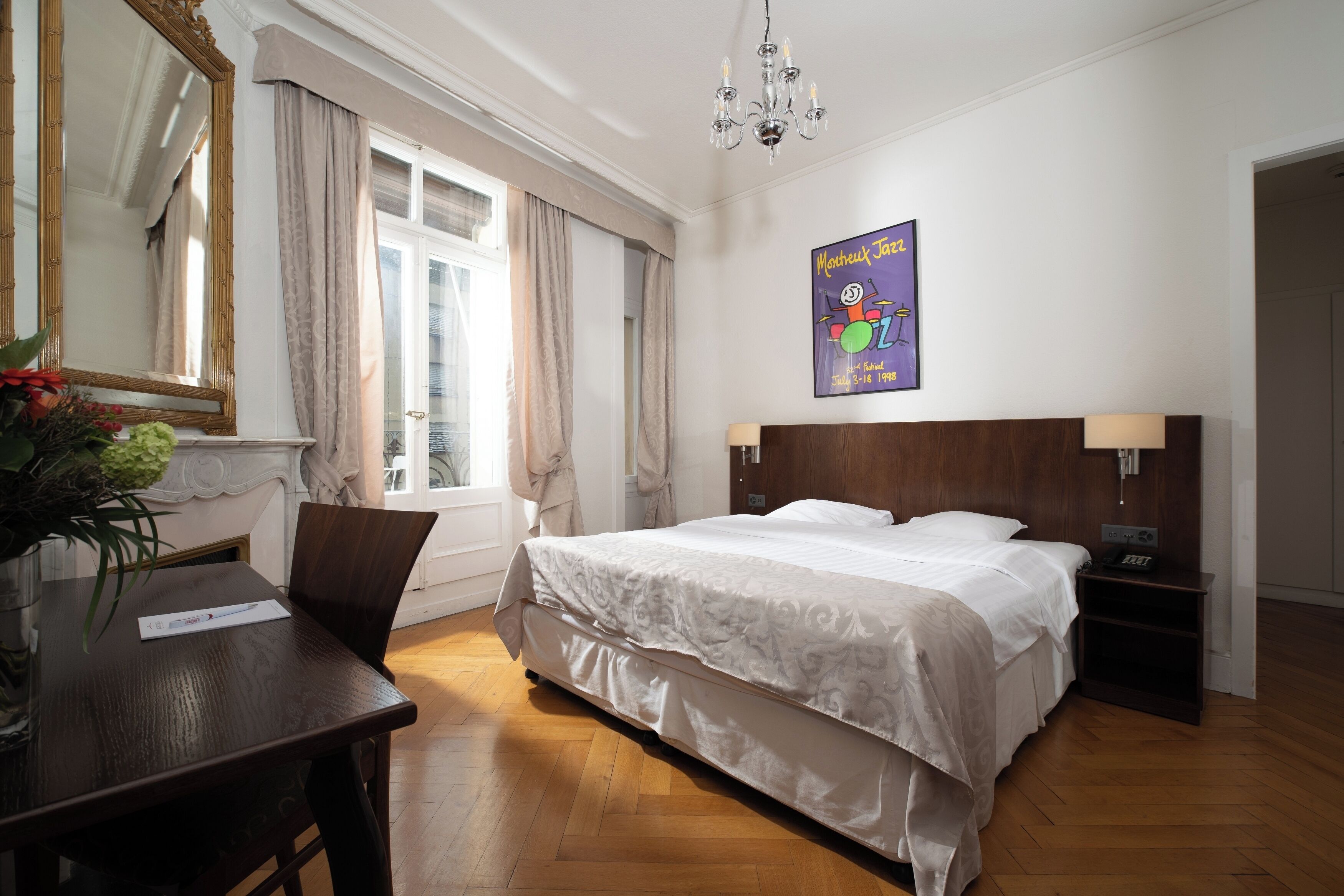 A hotel room with a large bed, a wooden nightstand, a desk with a chair, a chandelier, and a poster on the wall.