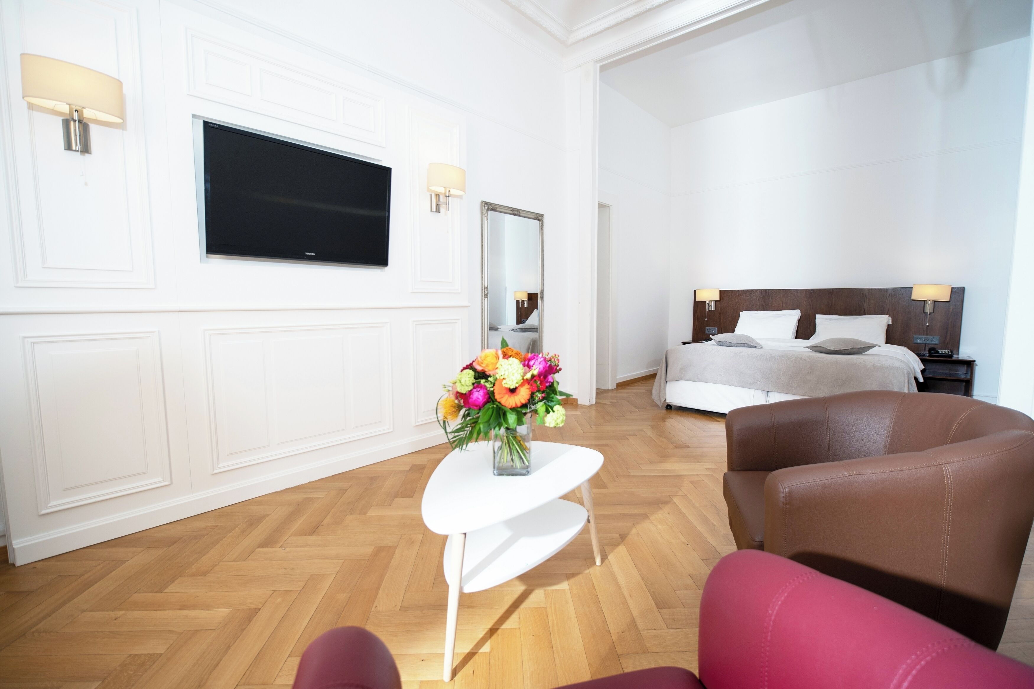 Une chambre d’hôtel avec un grand lit, un téléviseur fixé au mur, une petite table blanche ornée d’un vase de fleurs et un fauteuil en cuir marron.