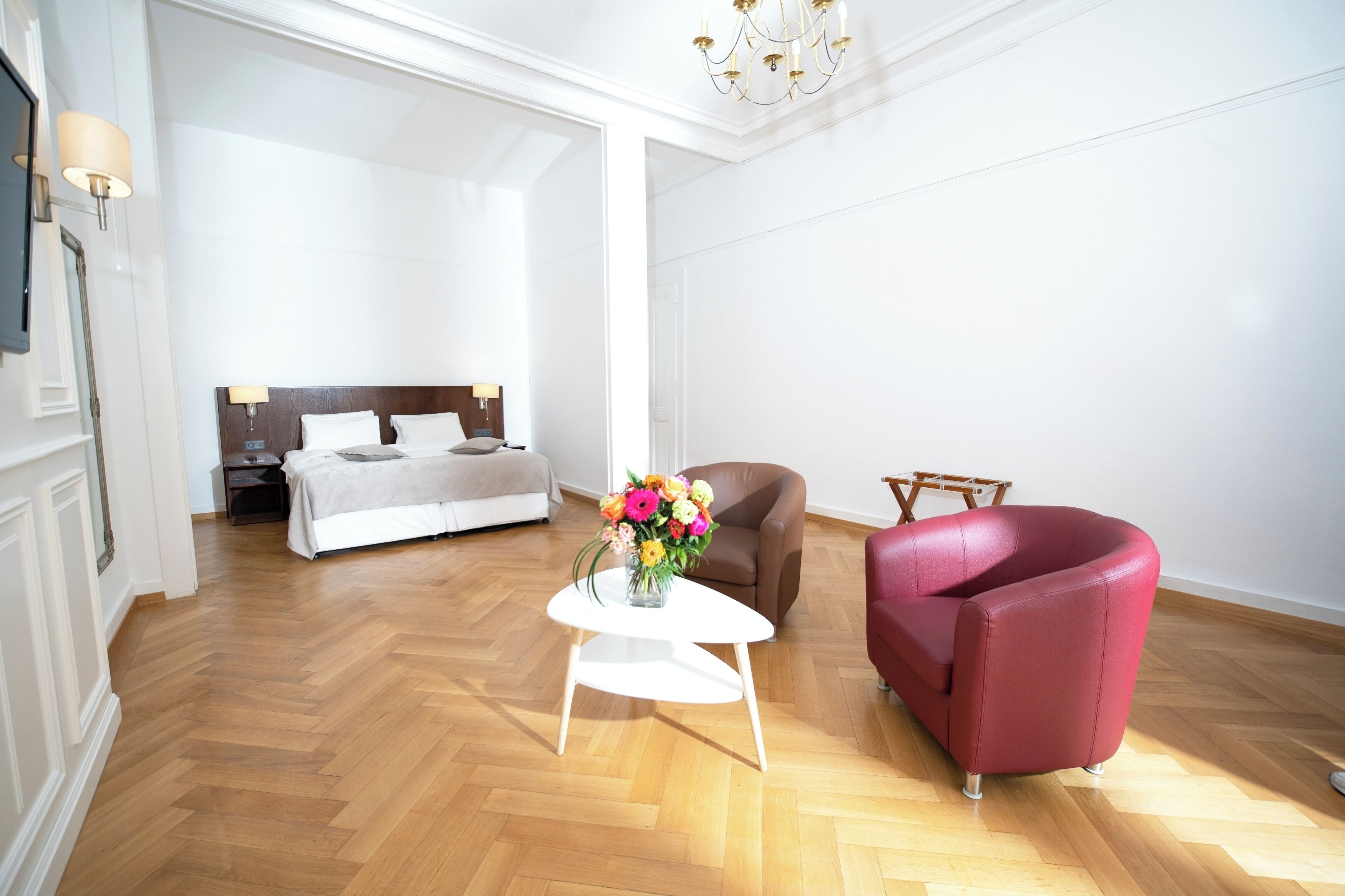 A hotel room with a wooden herringbone floor, a white bed with a wooden headboard, a brown armchair, a red armchair, and a white side table with a vase of flowers.