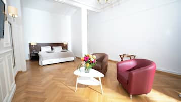 Quarto de hotel com piso de madeira em padrão herringbone, uma cama branca com cabeceira de madeira, uma poltrona marrom e uma poltrona vermelha, além de uma mesinha de cabeceira branca com um vaso de flores.