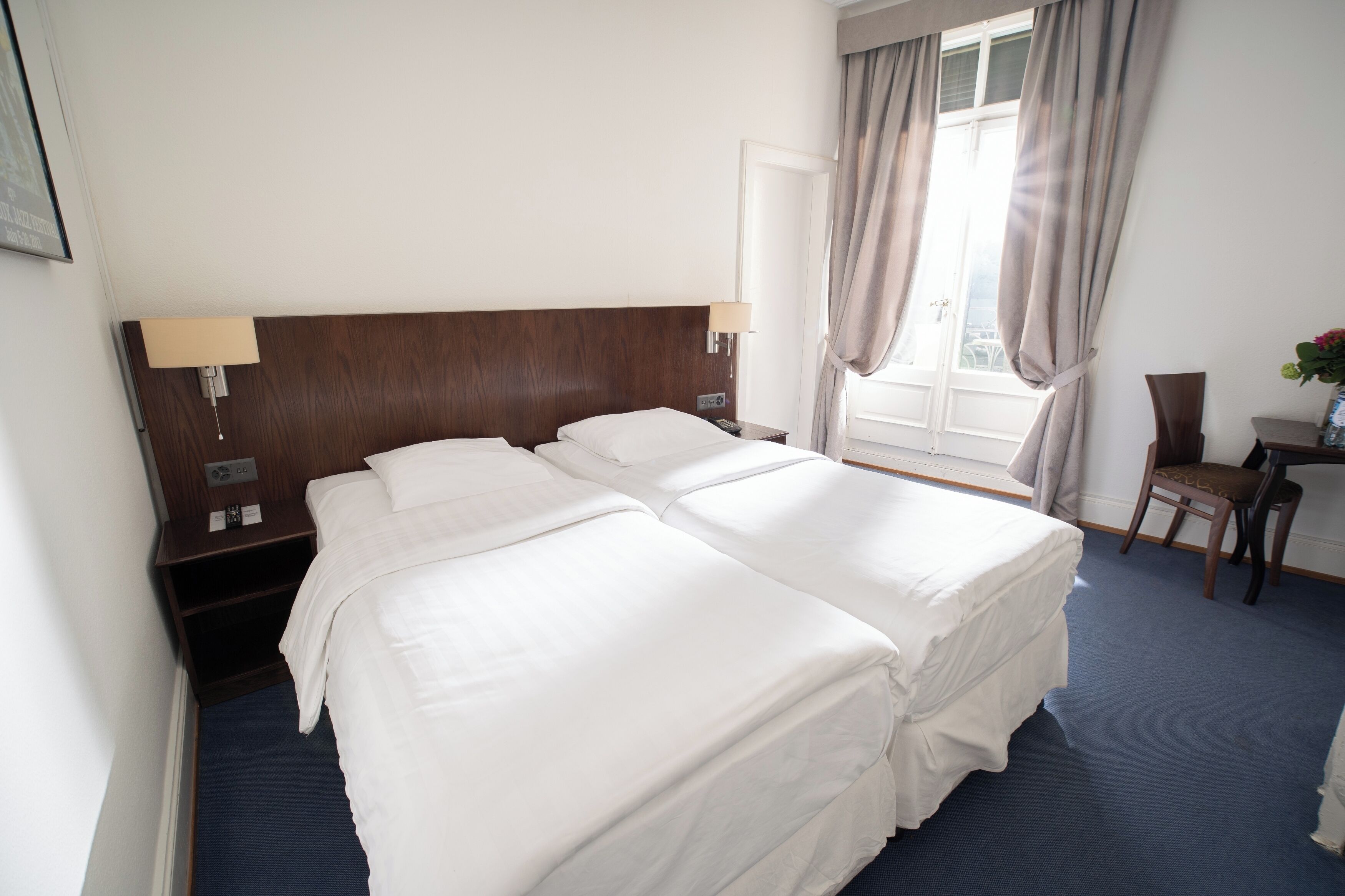 Habitación de hotel con dos camas, un escritorio, una silla, una ventana con cortinas y una mesita de noche con lámpara.