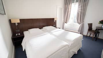 Habitación de hotel con dos camas, un escritorio, una silla, una ventana con cortinas y una mesita de noche con lámpara.