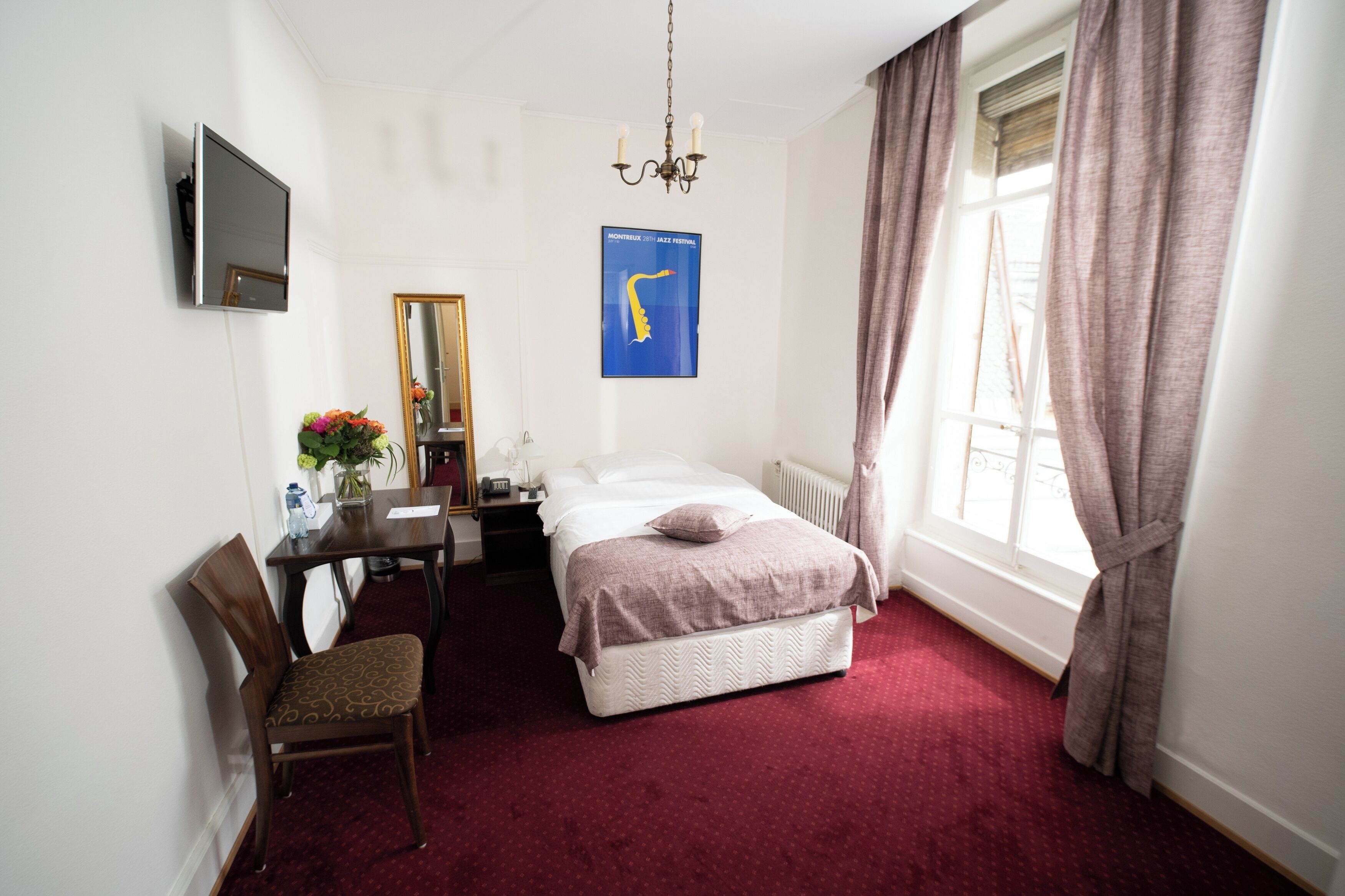 Une chambre d’hôtel équipée d’un lit, d’un bureau, d’une chaise, d’un miroir, d’une télévision et d’une fenêtre avec des rideaux.