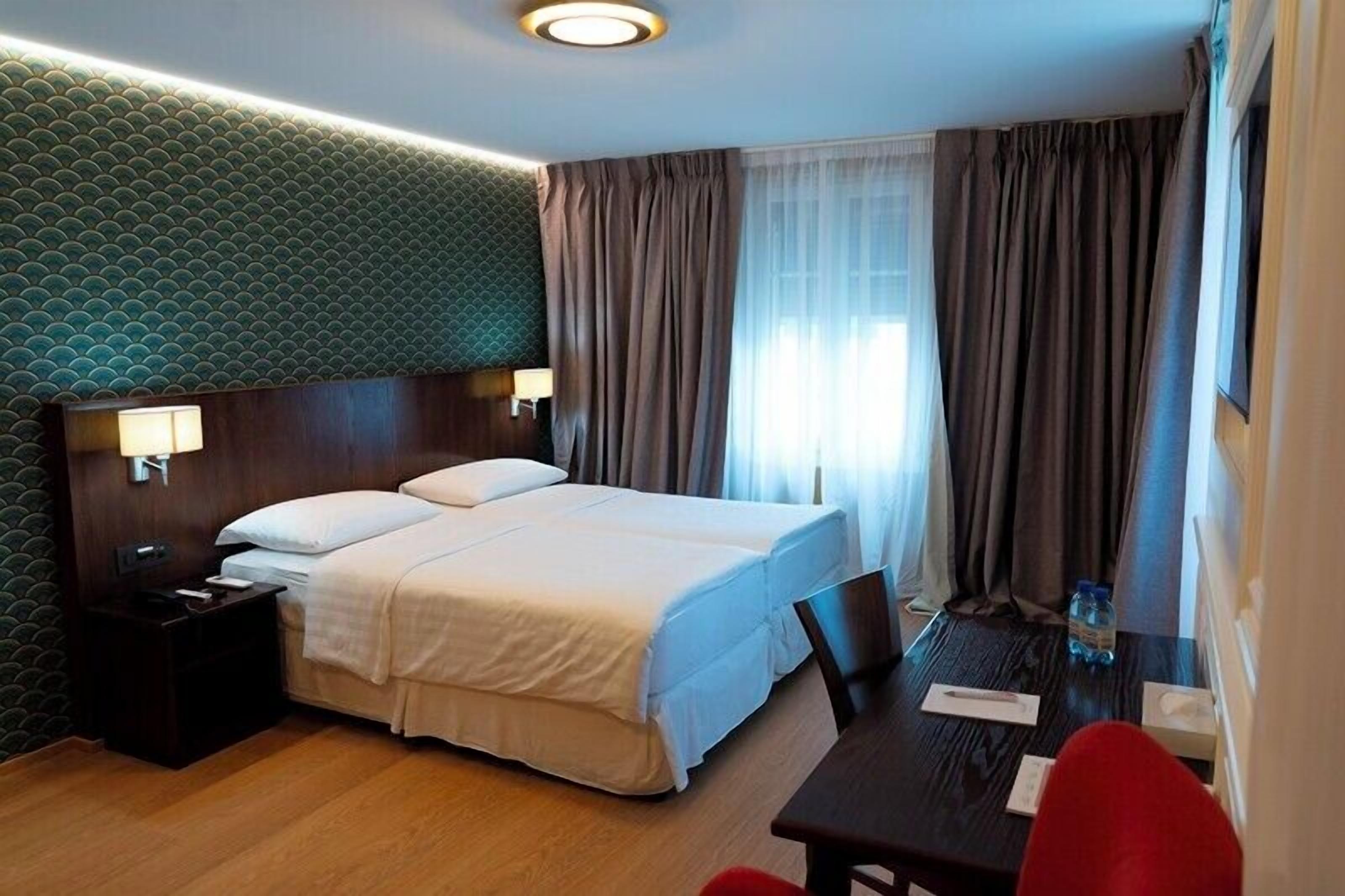 A hotel room with a double bed, a nightstand, a desk with a chair, and a window with curtains.