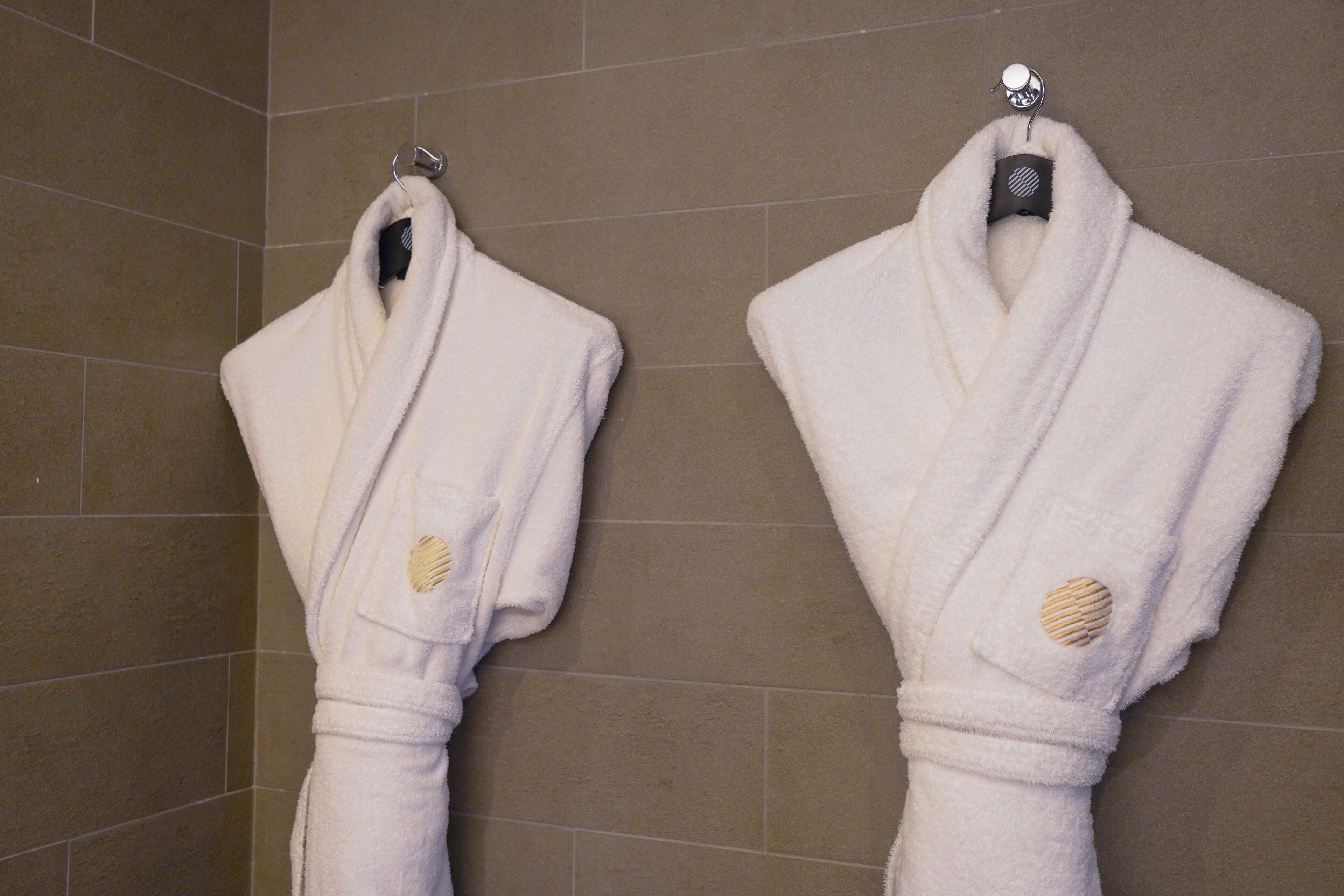 Deux peignoirs blancs sont suspendus à des crochets dans une salle de bain carrelée.