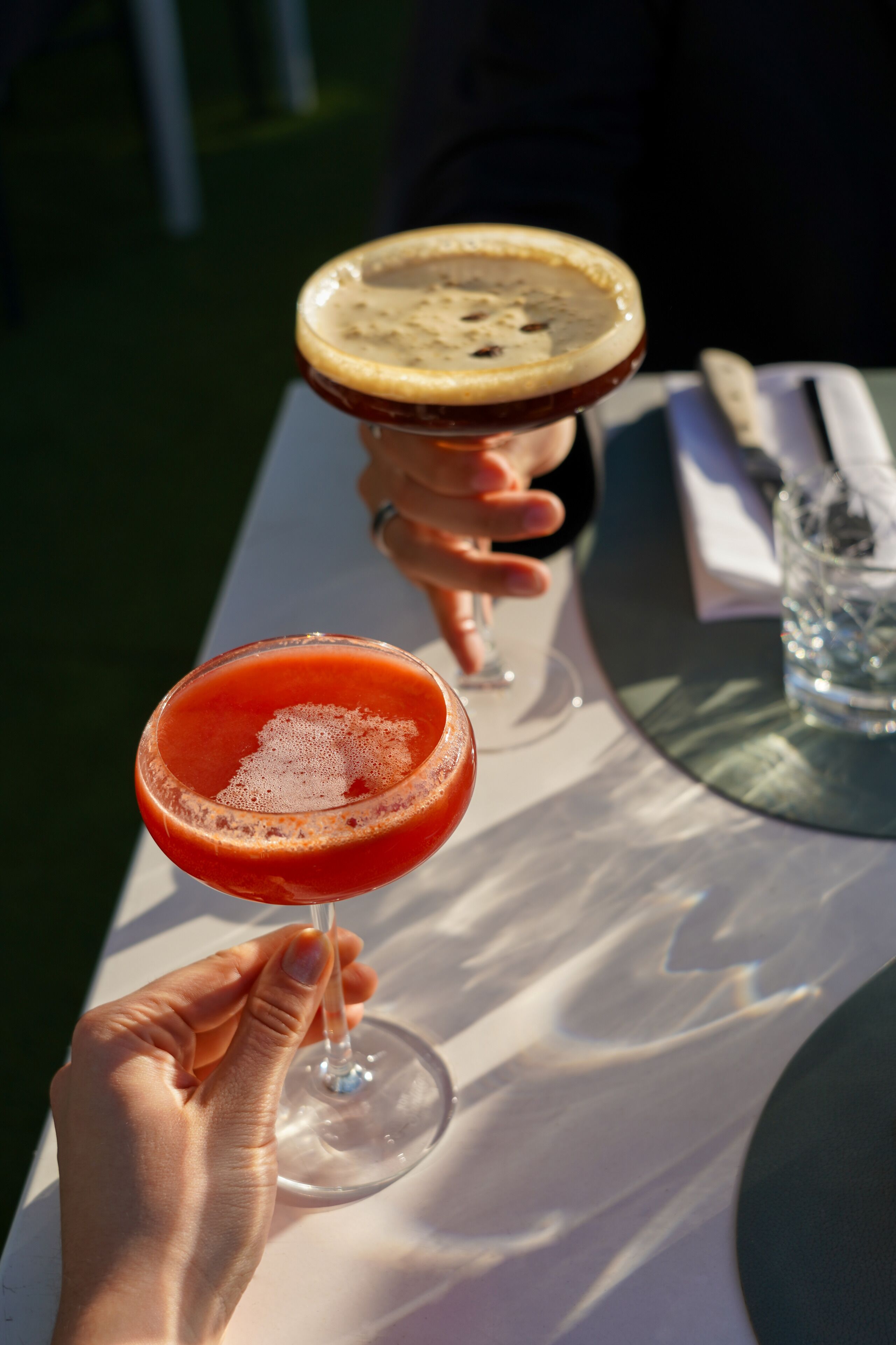 A hand holding a cocktail glass with a frothy drink, and another cocktail glass with a frothy drink in the background.