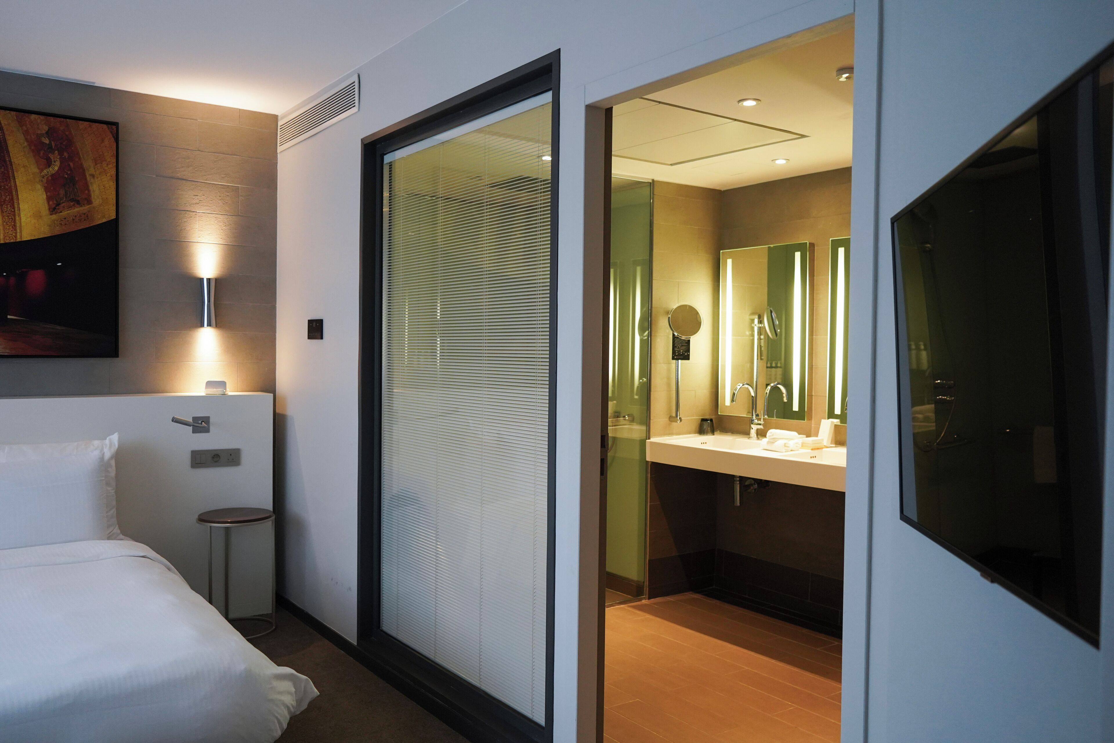Una habitación de hotel moderna con una cama, una pequeña mesa redonda, una lámpara de pared, un ventanal grande con persianas y un baño con lavabo y espejo.