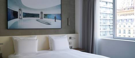 A hotel room with a large bed, a painting of a cityscape, a window with a view of buildings, and a curtain.